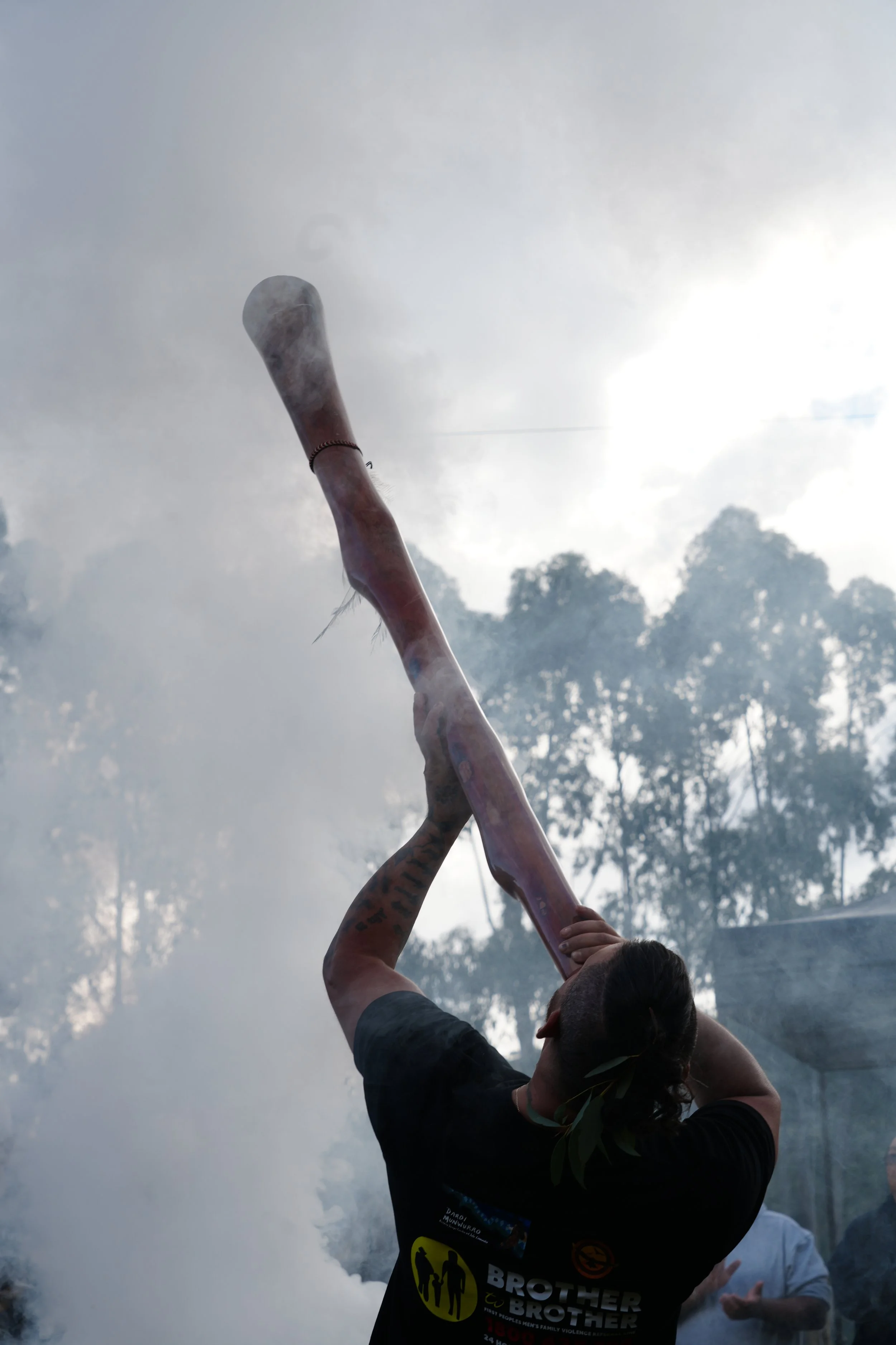 A person holding a burning torch with smoke and haze in the background, trees and other people are also visible.