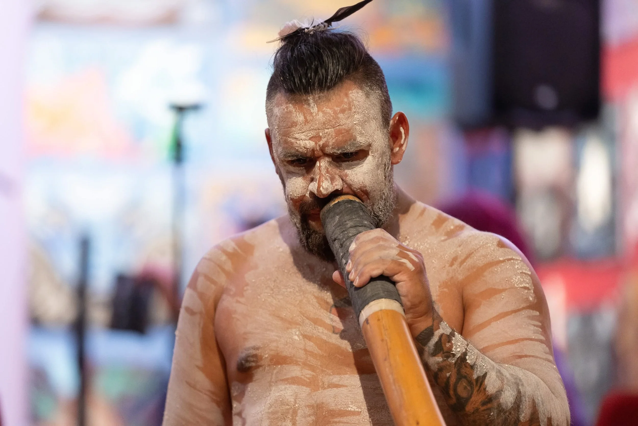 A shirtless man with clay painted on his face and body holding a traditional didgeridoo, with colorful blurred background.