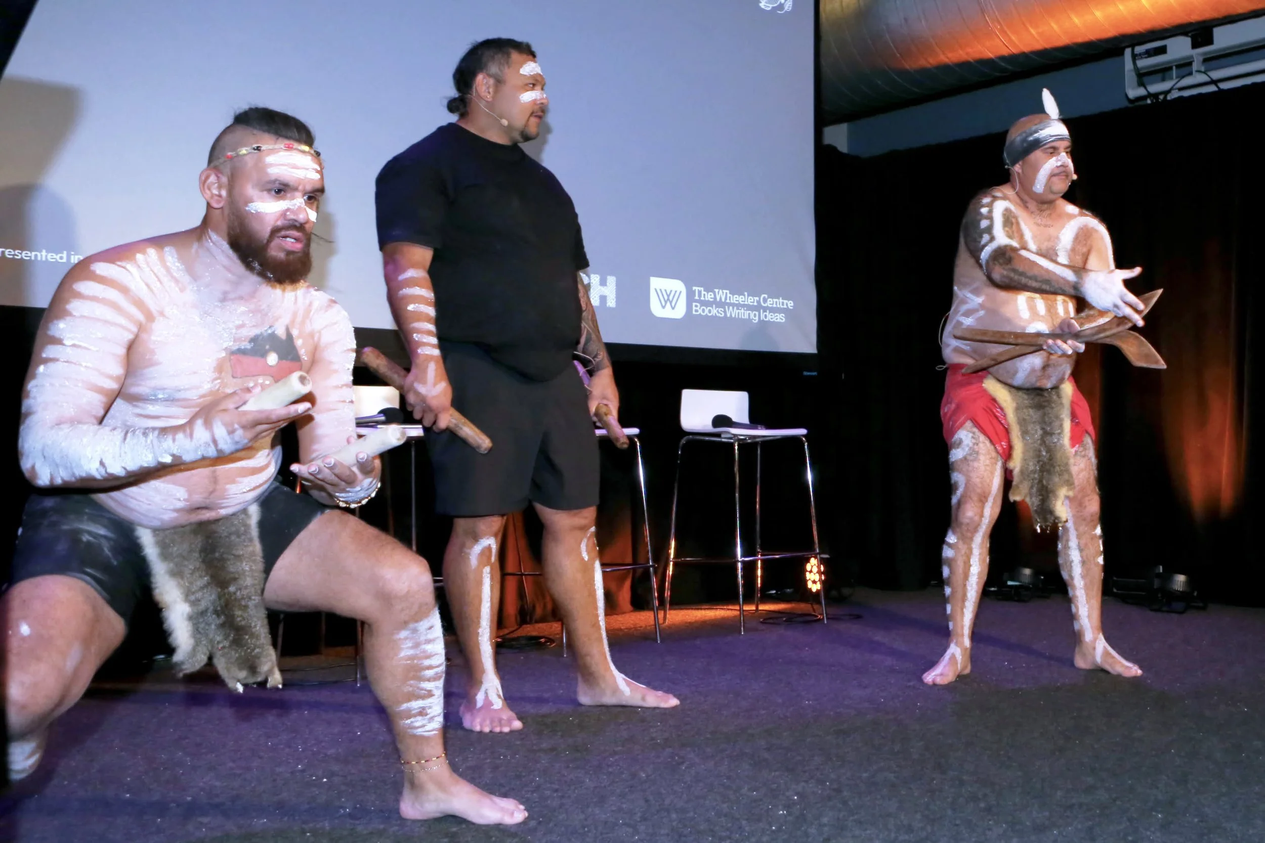 Two men and one woman dressed in Indigenous Australian body paint and traditional attire on stage, with a man in the middle observing, possibly in a cultural performance or presentation.