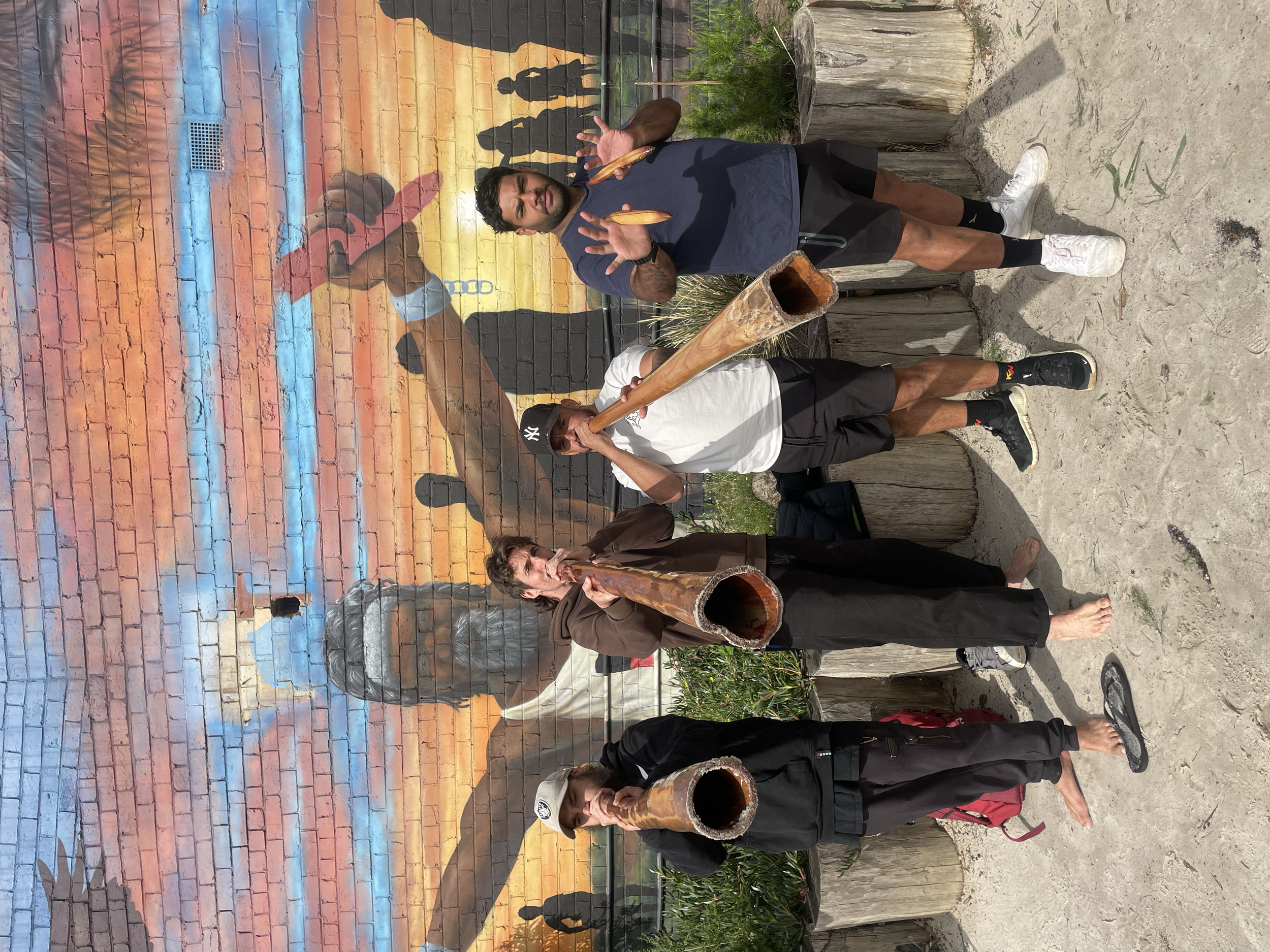 Four men standing outdoors in front of a colorful mural on a brick wall, playing didgeridoos. The mural depicts a man with outstretched arms, a sunset sky, and silhouette figures, with some elements of street art and graffiti.