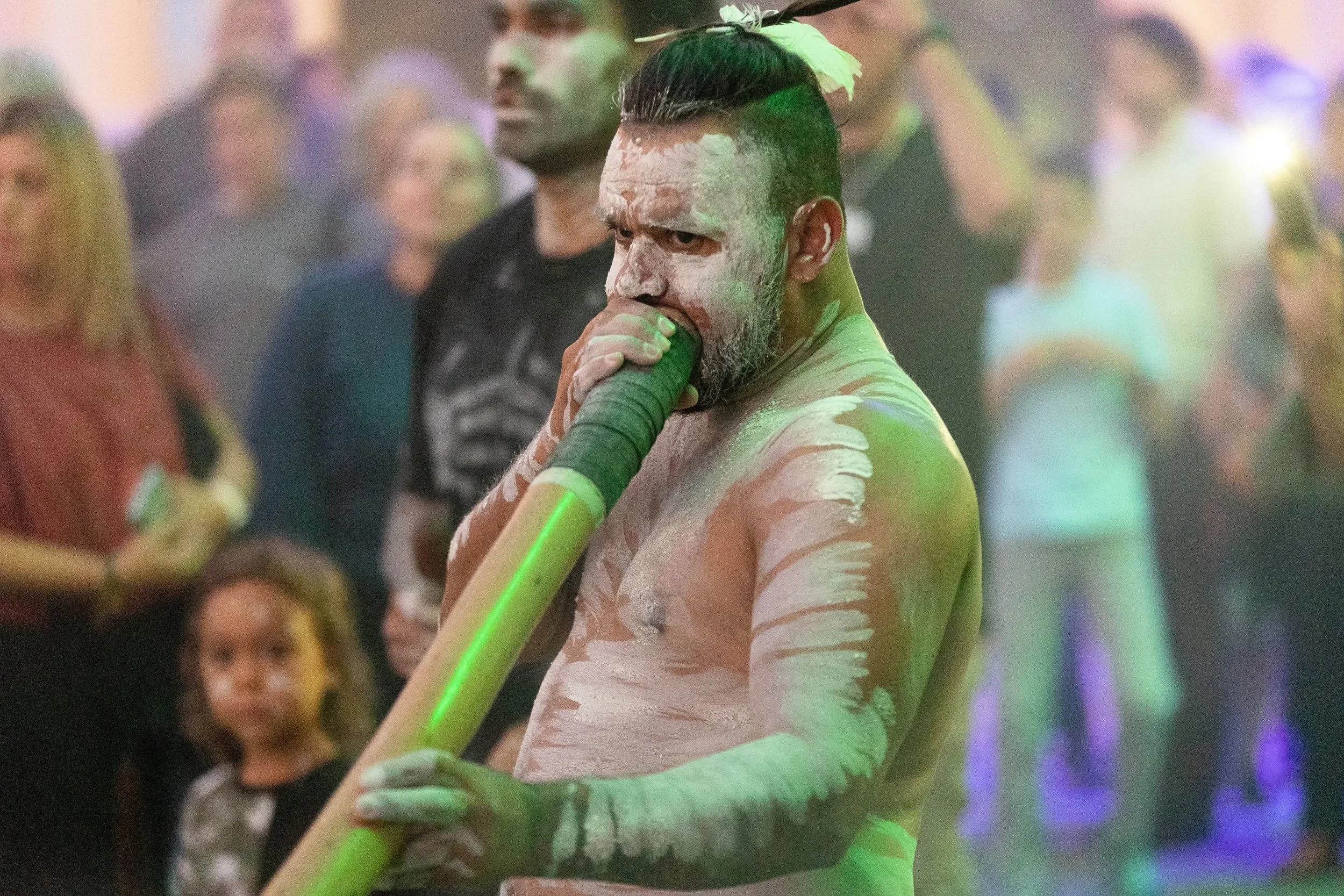 A man with face paint and body paint in light colors, holding a green glow stick, at a party or festival with blurred people in the background.