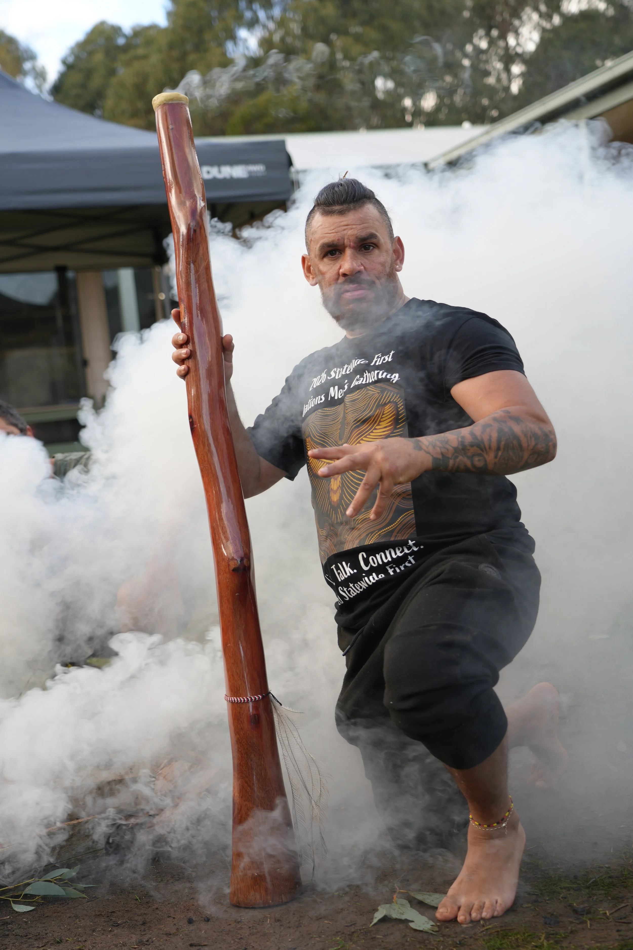 A man crouches in smoke, holding a large, carved wooden stick, with a serious expression.
