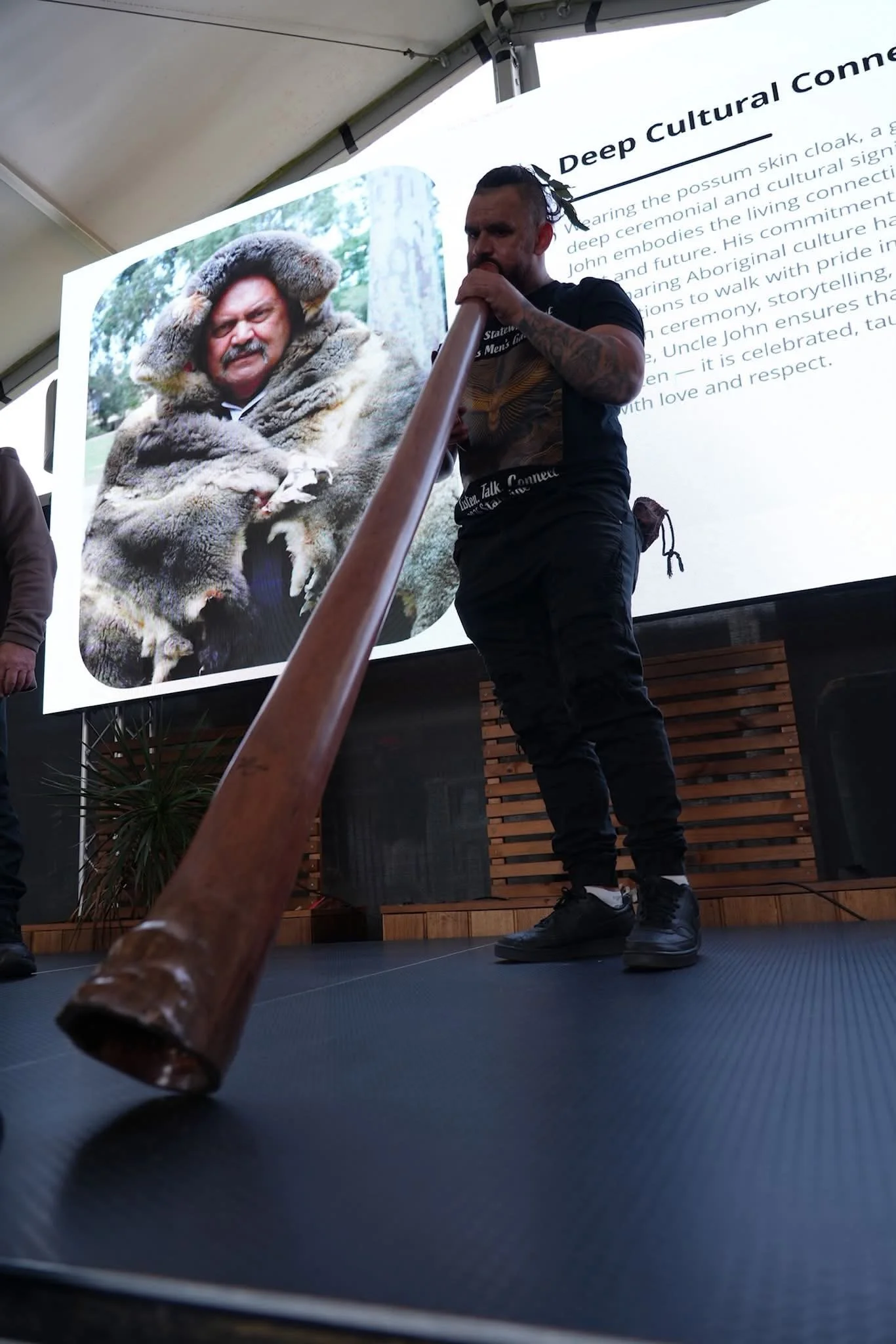 A man playing a didgeridoo on stage during a presentation with a large screen behind him. The screen displays a photo of a man in traditional Indigenous Australian attire and some text about deep cultural connections.