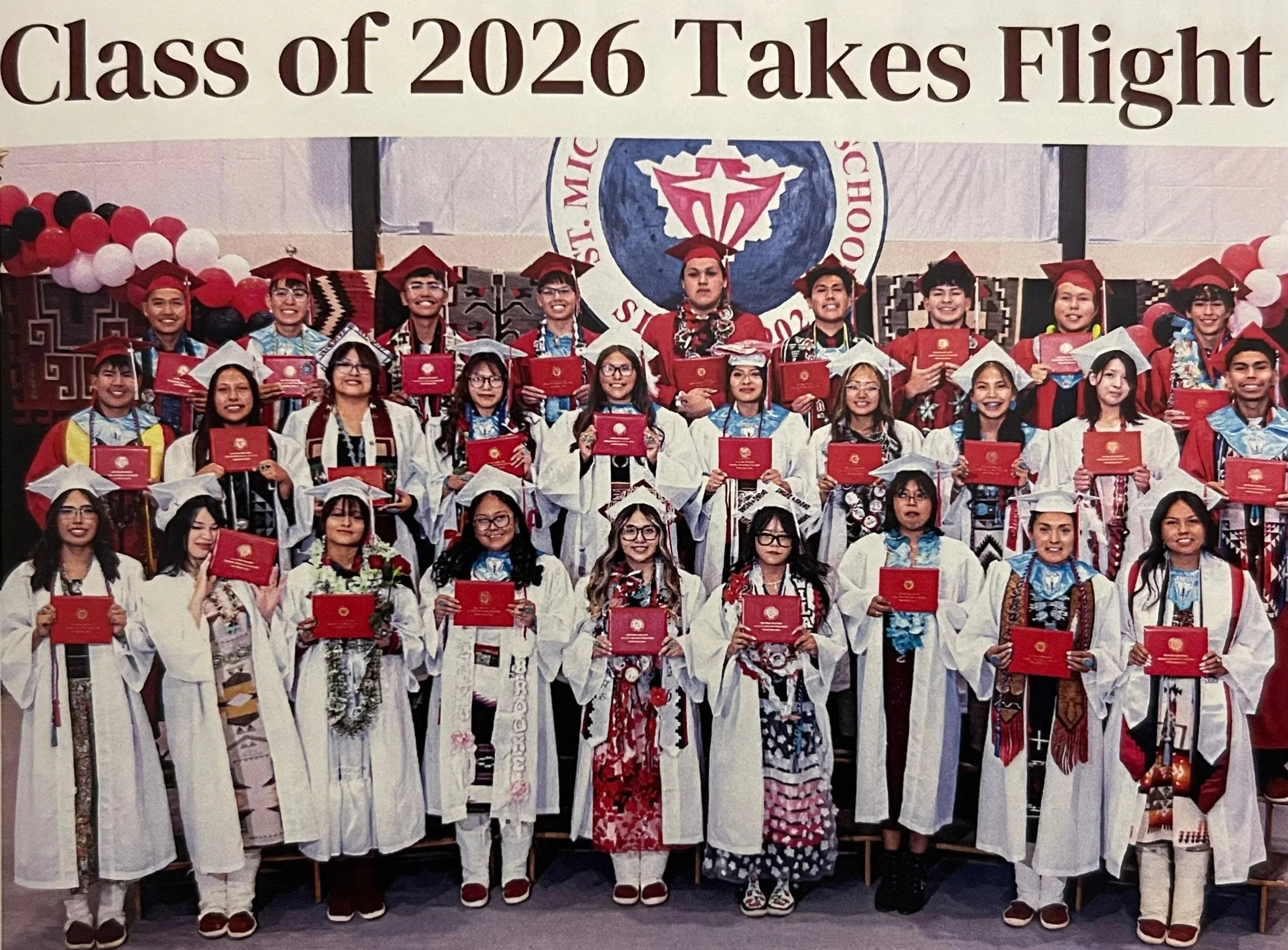 Group of graduates in traditional attire holding red diplomas during a graduation ceremony. A large emblem with a diamond shape is visible in the background, along with colorful balloons.
