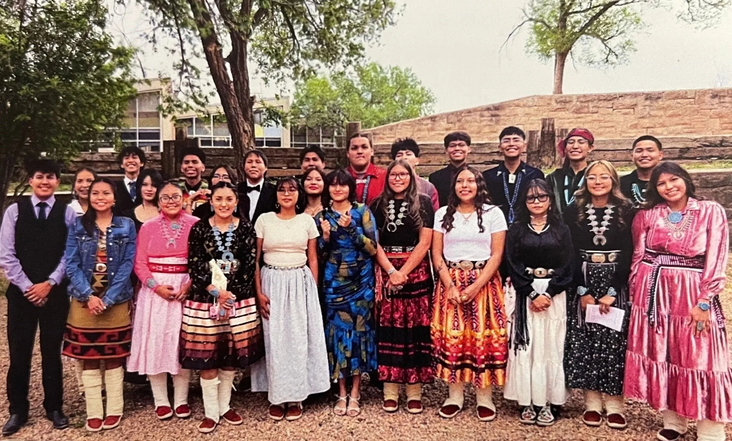Group of people dressed in traditional clothing outdoors near trees and a brick wall.