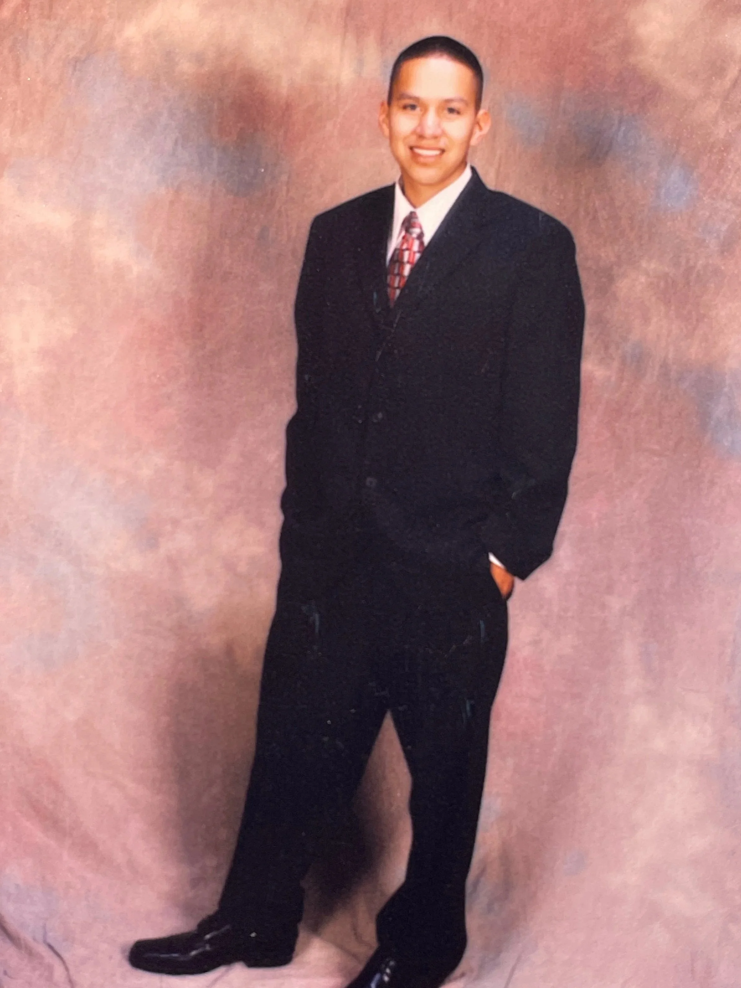 A young man in a dark suit, white shirt, and colorful tie, standing indoors in front of a textured pink wall, smiling for the camera.