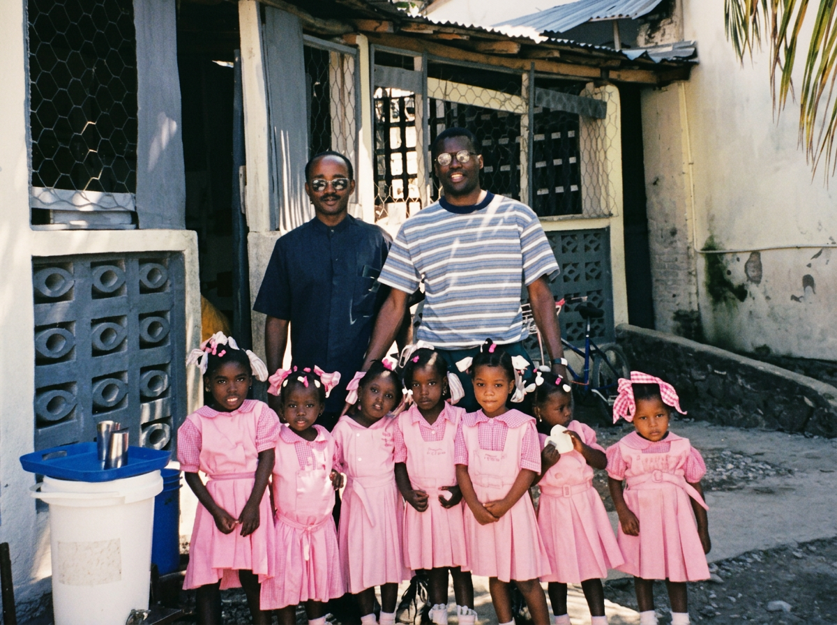 Two men, one in a striped shirt and the other in a dark button-up, standing behind six young girls dressed in pink uniforms with bows in their hair. They are outdoors in front of a building with a concrete wall and a metal fence.