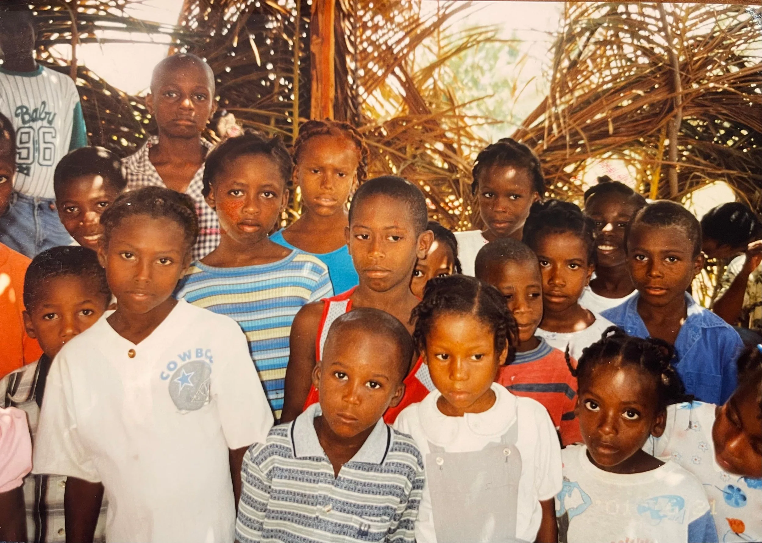 A group of children and young people standing together inside a structure made of palm leaves, looking at the camera.