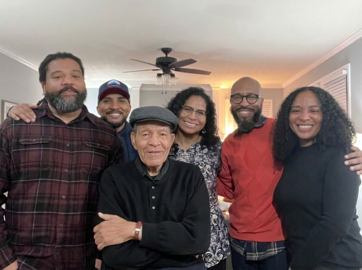 Six people, three men and three women of diverse ages, smiling and gathered together in a living room.