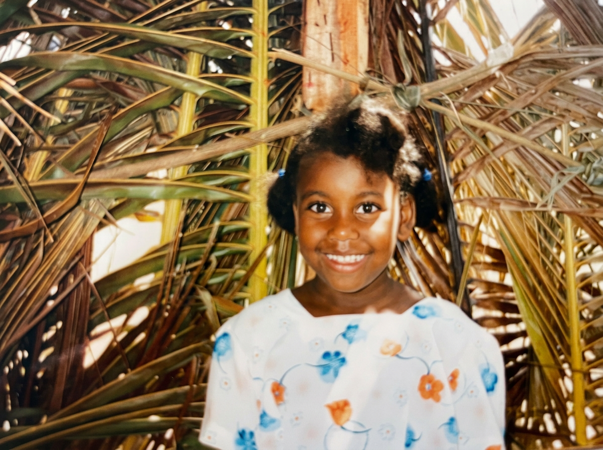A young girl with a big smile, wearing a white dress with orange and blue floral patterns, standing in front of tropical foliage with palms and banana leaves.
