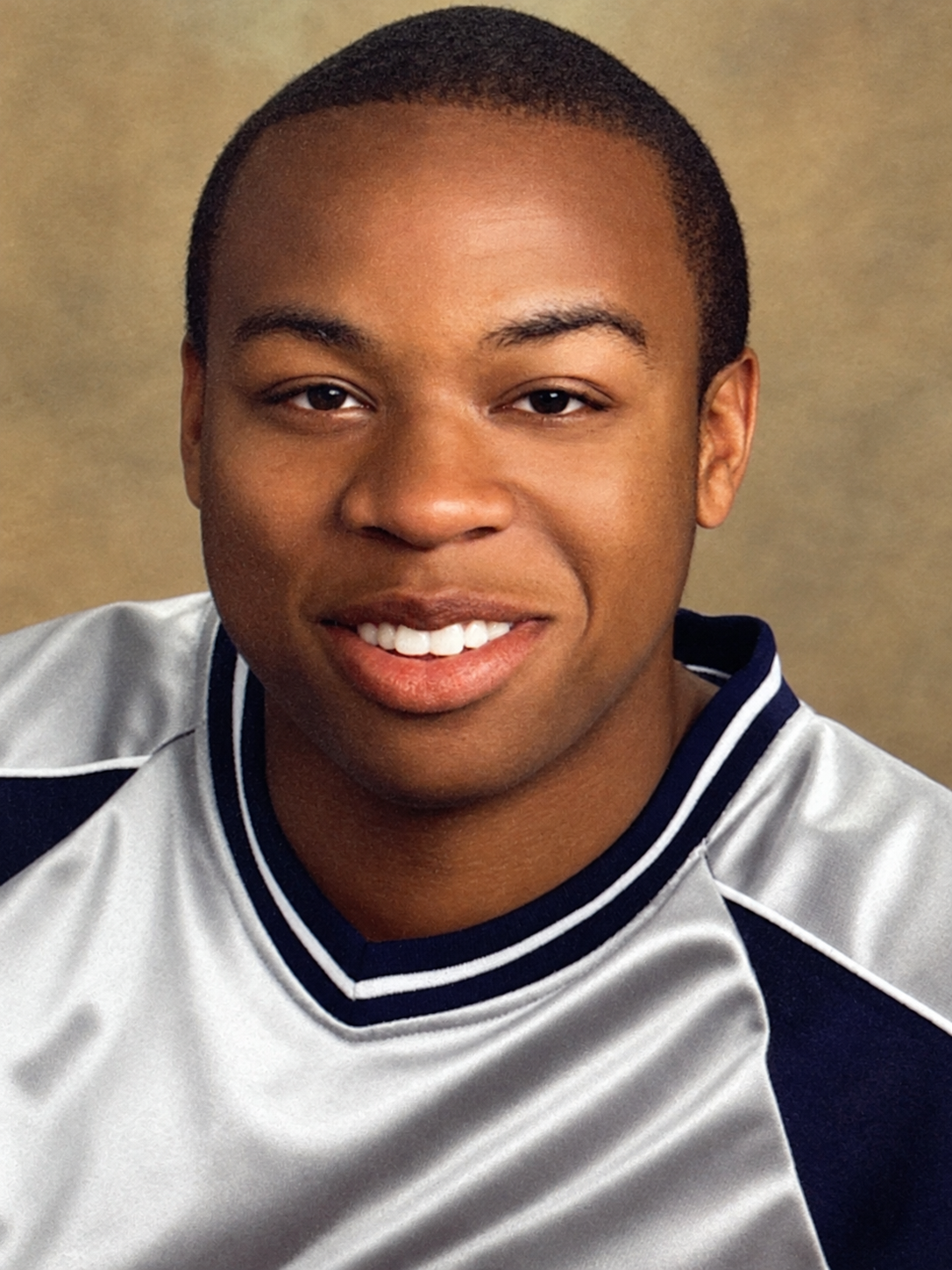 Close-up portrait of a young man with short hair, smiling, wearing a white and navy sports jersey, against a neutral background.