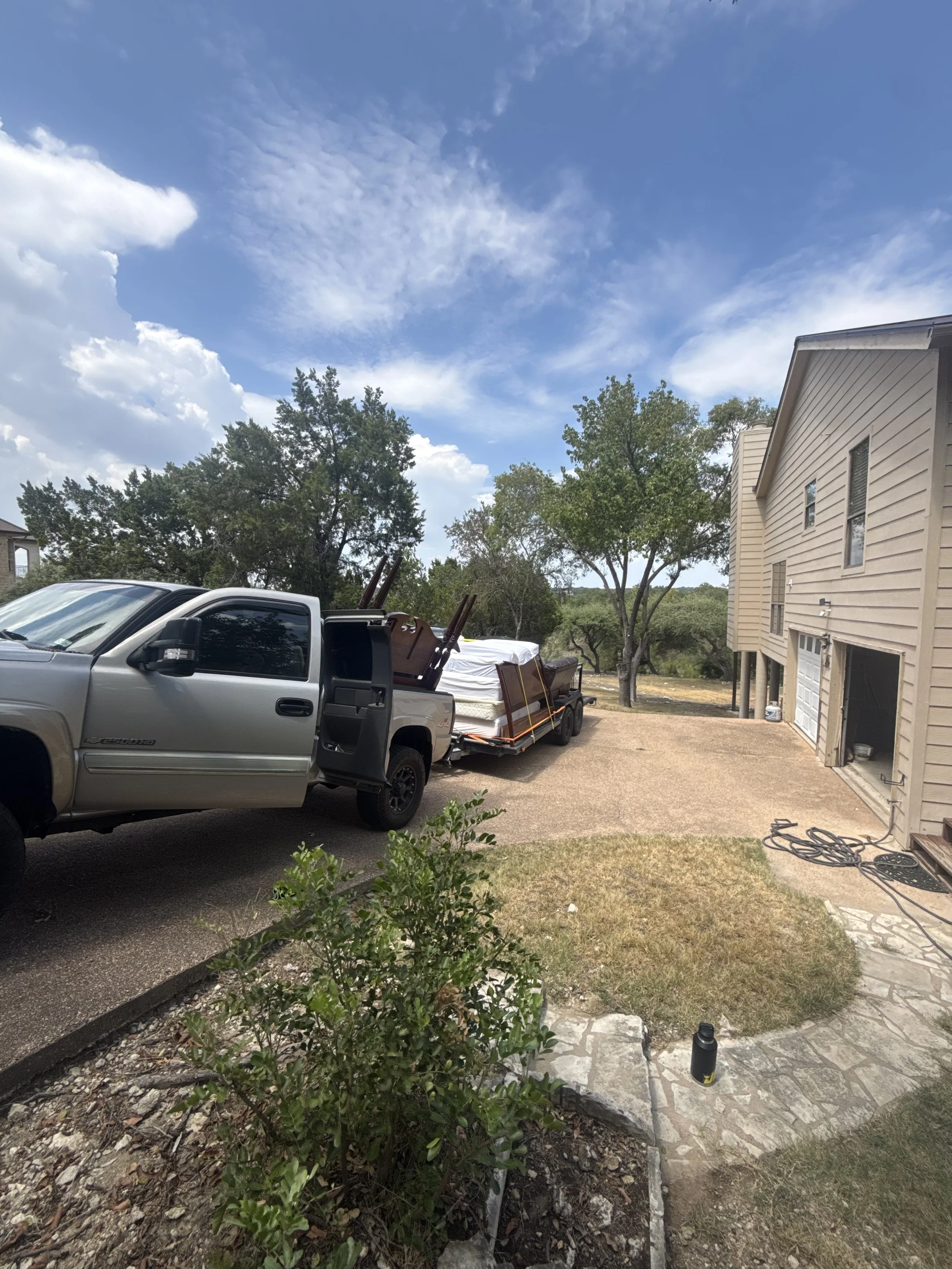 View of a house with a driveway and a pickup truck pulling a trailer loaded with furniture and supplies, surrounded by trees and cloudy sky.