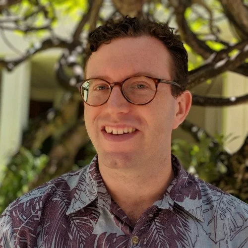 A smiling man with glasses in front of a tree and a house.
