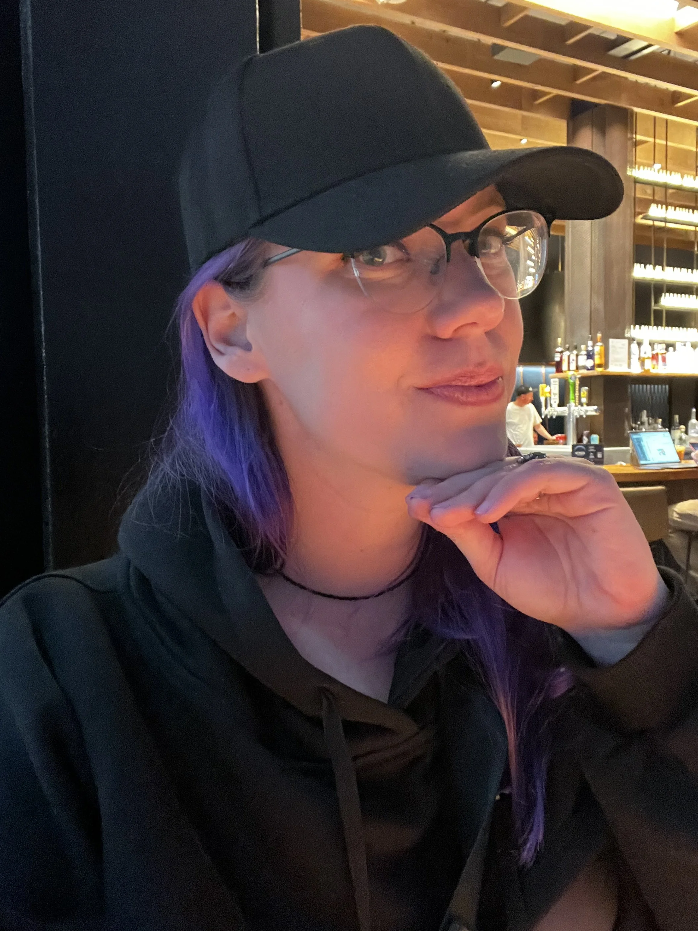 A person with purple hair wearing a black baseball cap, glasses, and a black hoodie, sitting at a bar or restaurant with a dimly lit background that includes bottles and bar equipment.