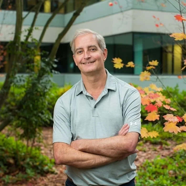 A middle-aged man with gray hair standing outdoors with arms crossed, smiling, in front of a building and colorful trees with red, yellow, and green leaves.
