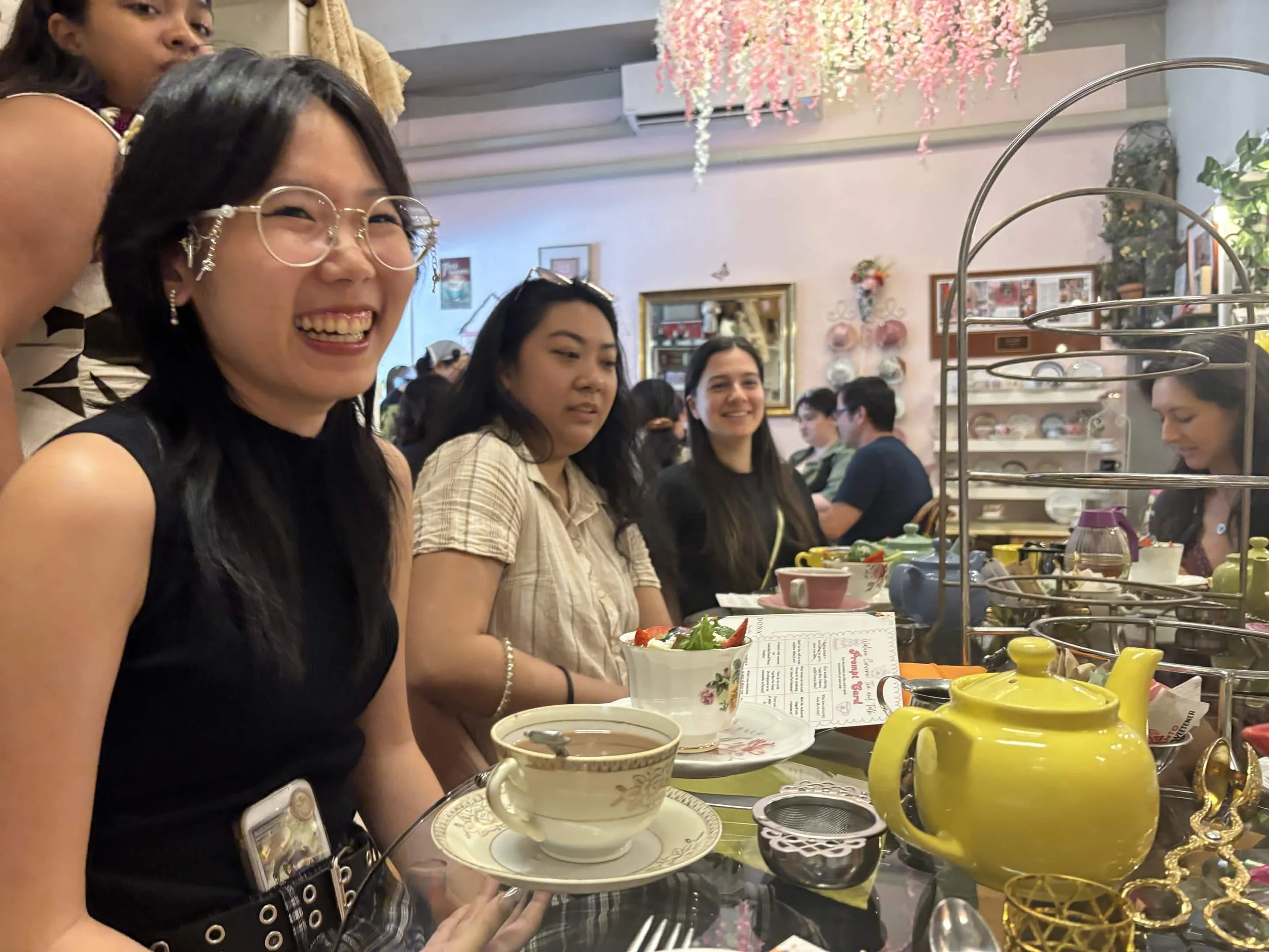 Group of women seated at a table in a restaurant, enjoying tea and dessert, with a decorative display of yellow teapots and teacups in the foreground.