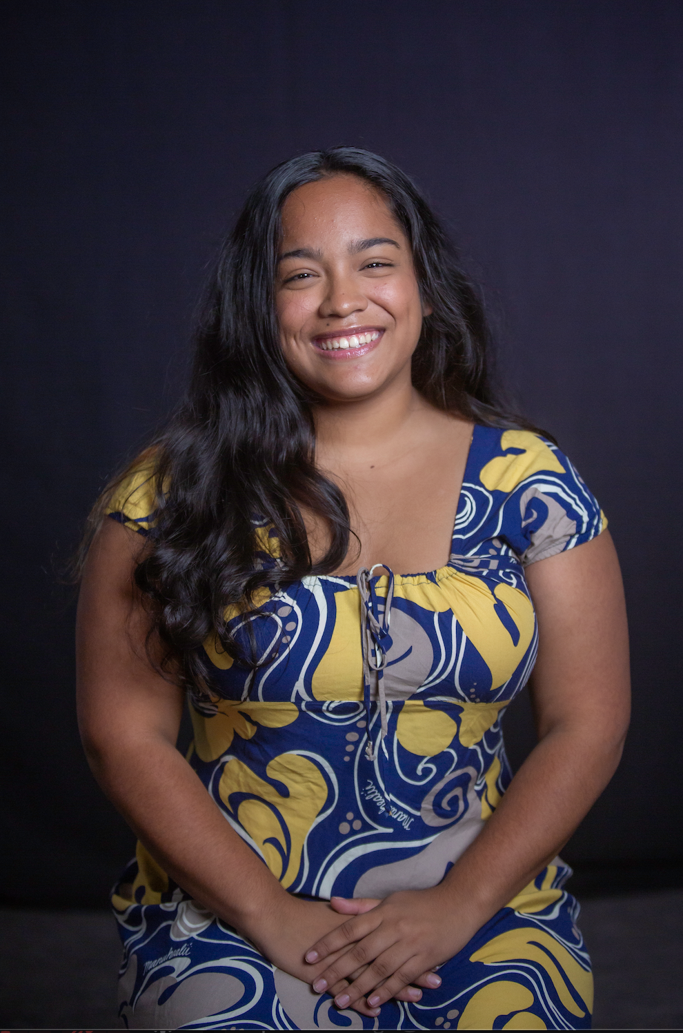 A young woman with long dark curly hair smiling while sitting against a dark background, wearing a colorful yellow, blue, and beige patterned dress.