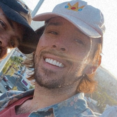 Two men smiling outdoors, one wearing a baseball cap with a crown logo, the other wearing a dark cap, sunlight shining on them.