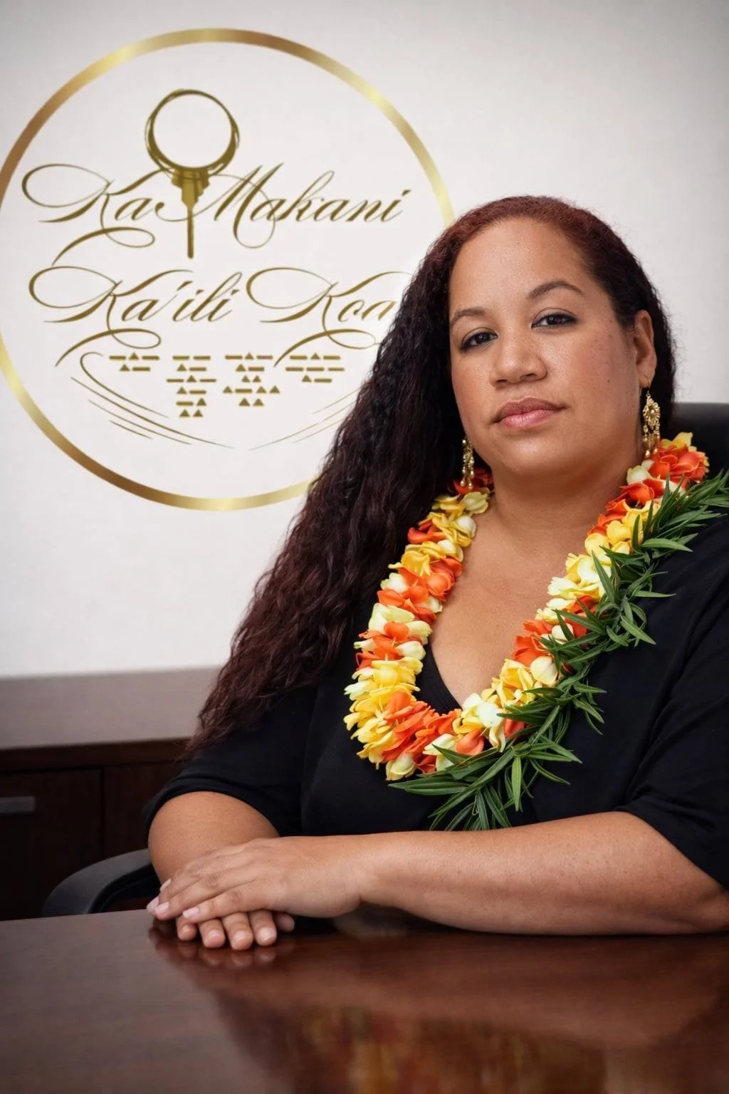 A woman with long, curly hair wearing a black top, gold earrings, and a colorful leis necklace sitting at a desk with a logo behind her that includes a magnifying glass and text in a script font.