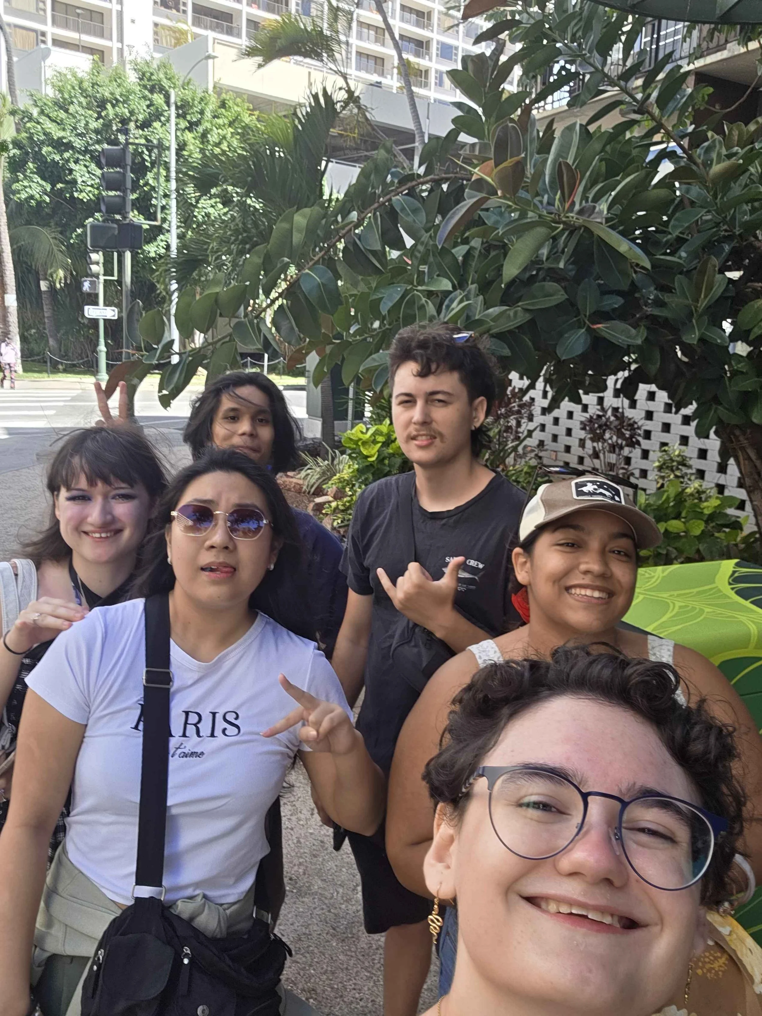 A group of diverse people posing for a selfie outdoors in an urban area, with greenery and tall buildings in the background.