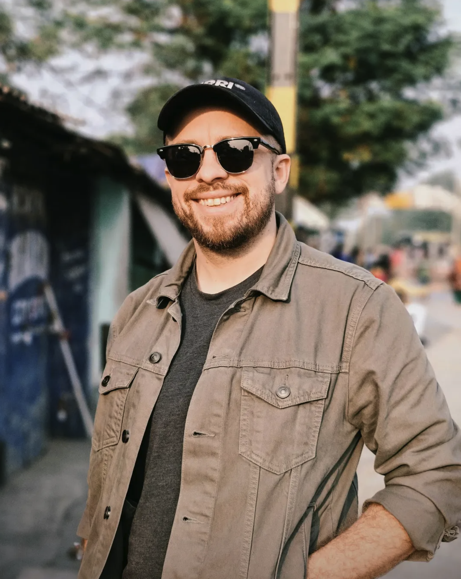 A smiling man wearing sunglasses and a black baseball cap, with a beard, in an outdoor setting surrounded by trees and blurred background.