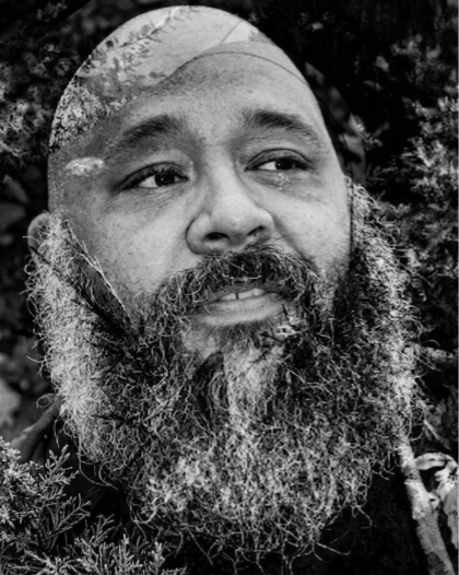 Close-up black and white photo of a man with a beard and mustache, outdoors with trees in the background.