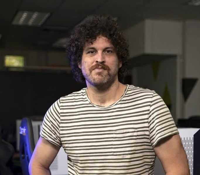 A man with curly hair and a beard wearing a striped T-shirt, standing indoors in a casual setting.