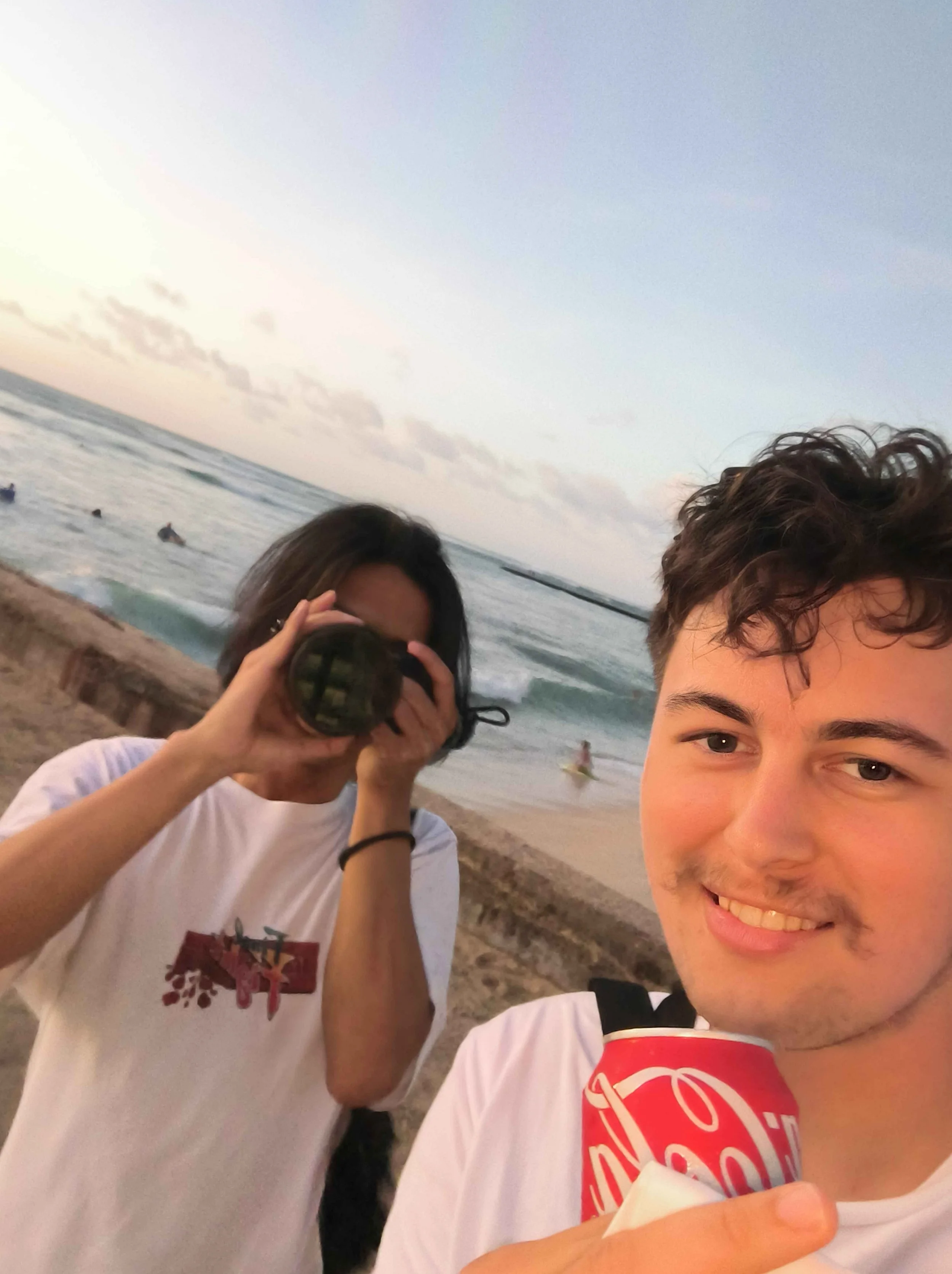 Two young men at the beach, one holding a camera and the other holding a Coca-Cola can, smiling with ocean and sunset in the background.