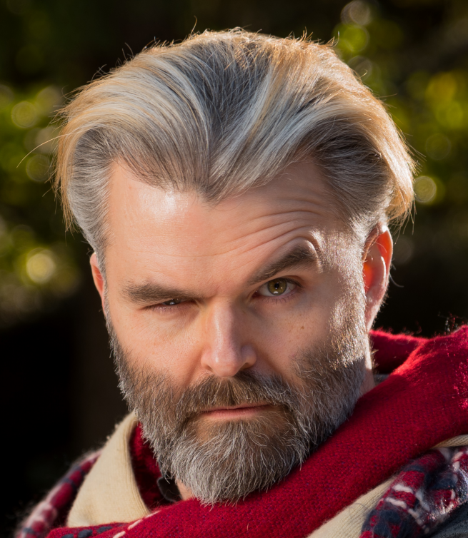 A man with salt-and-pepper hair and beard, winking with one eye, outdoors with a blurred background of trees or bushes.