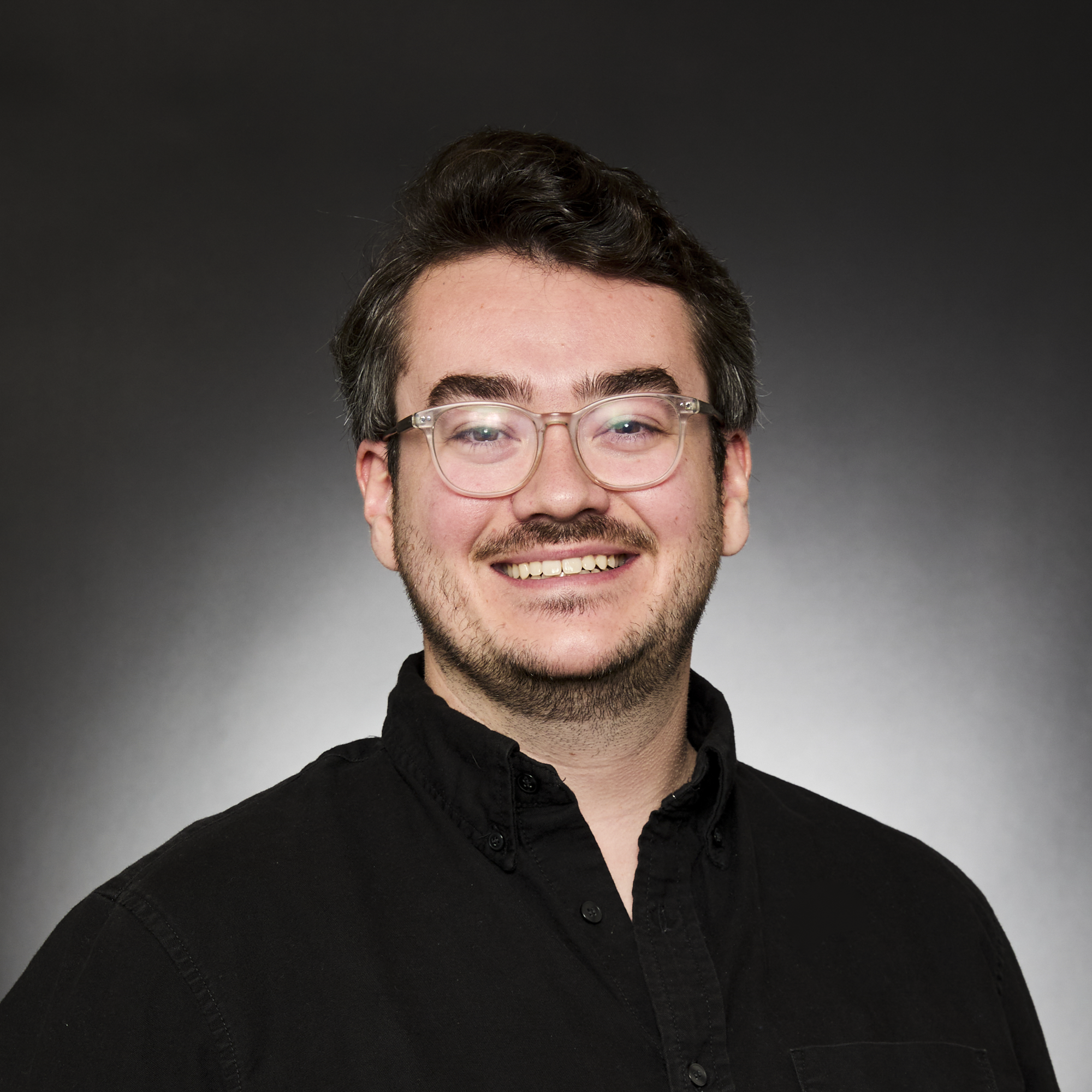 Close-up of a man's face with curly dark hair, clear glasses, and a slight smile, wearing a black shirt against a dark gradient background.