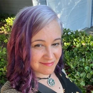 A woman with purple and pink hair smiling outdoors near green bushes and a building.
