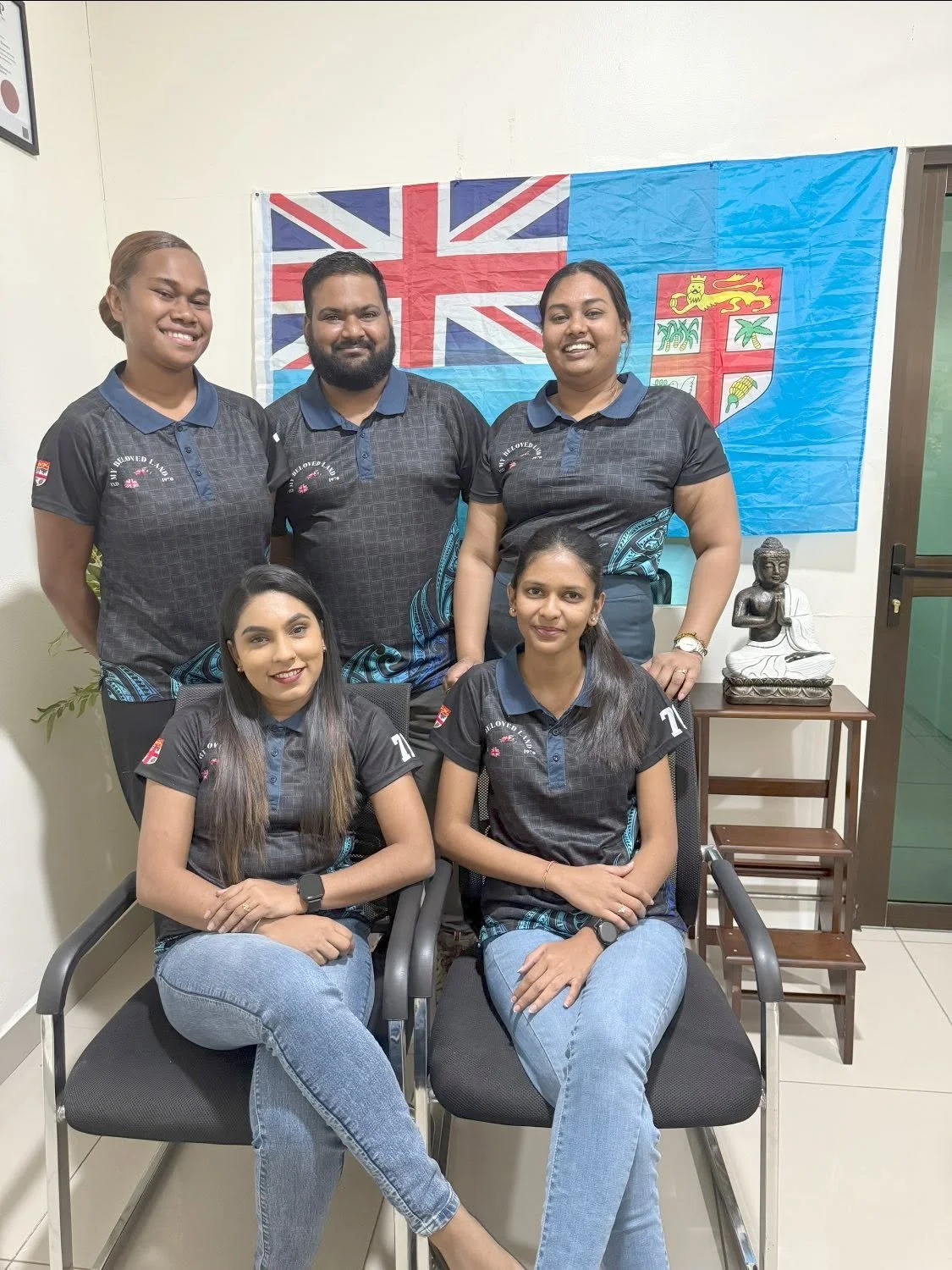 Group photo of five people wearing matching black and blue shirts, with a flag featuring the Union Jack and a blue background in the background.