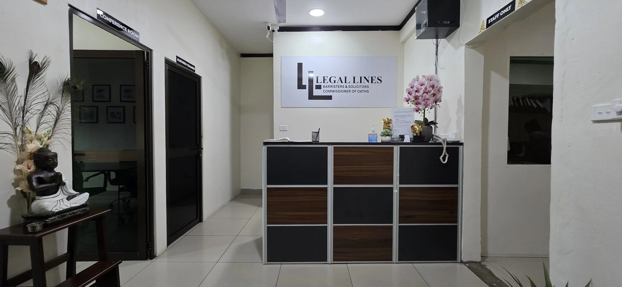 Reception area of a law office with a sign that reads 'Legal Lines, Barristers & Solicitors, Commissioner of Oaths.' There is a wooden divider in front of the reception desk. Decor includes a potted orchid and gold animal figurines on the counter. To the left, there is a small statue of a seated meditating figure and a peacock feather arrangement. Two doors labeled 'Conference Room' and 'Partner's Chamber' are on the left, and a window opening to another room is on the right.
