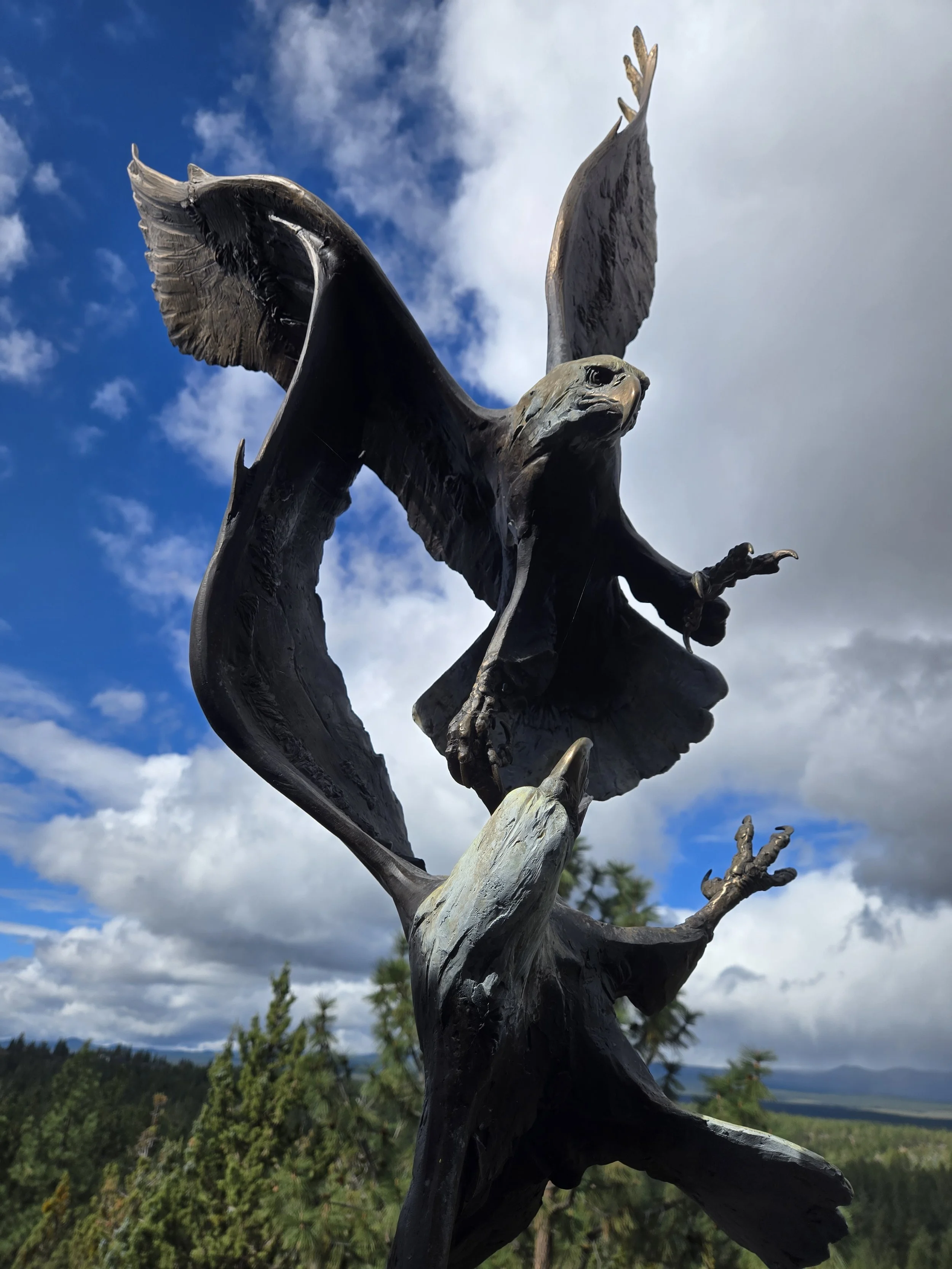 Bronze statue of a bird with wings spread, perched on a branch, against a scenic background of blue sky and clouds with distant trees and mountains.