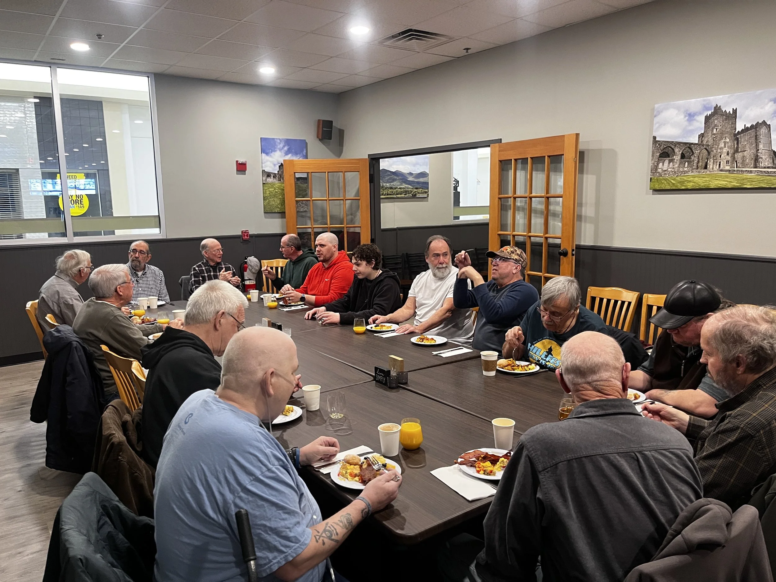 People gathered around a large table in a restaurant, eating breakfast or brunch, with Mountain Dew and juice glasses on the table. The restaurant has gray walls, wooden doors, landscape photographs, and a large window showing outside view.
