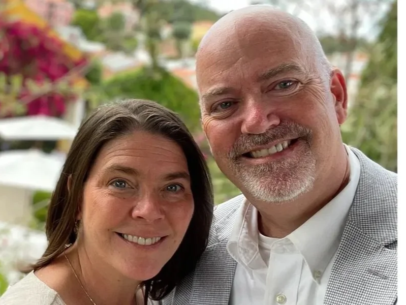 A smiling woman with shoulder-length brown hair and a man with a bald head and a white beard, both dressed in semi-formal attire, standing outdoors with a blurred background of greenery and colorful flowers.