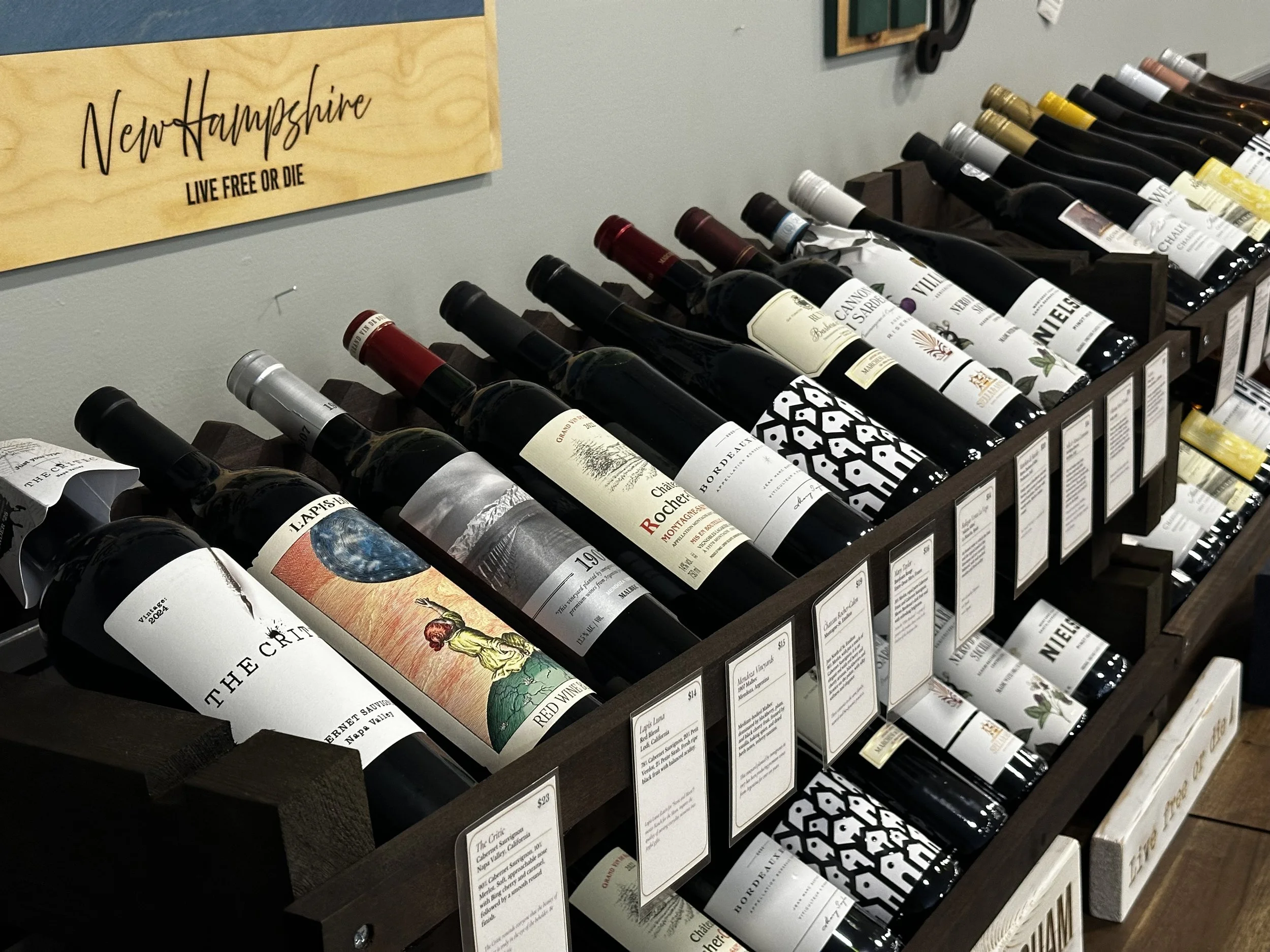 Display of wine bottles arranged in a wooden rack with labels visible. The background features a sign that reads 'New Hampshire Live Free or Die'.