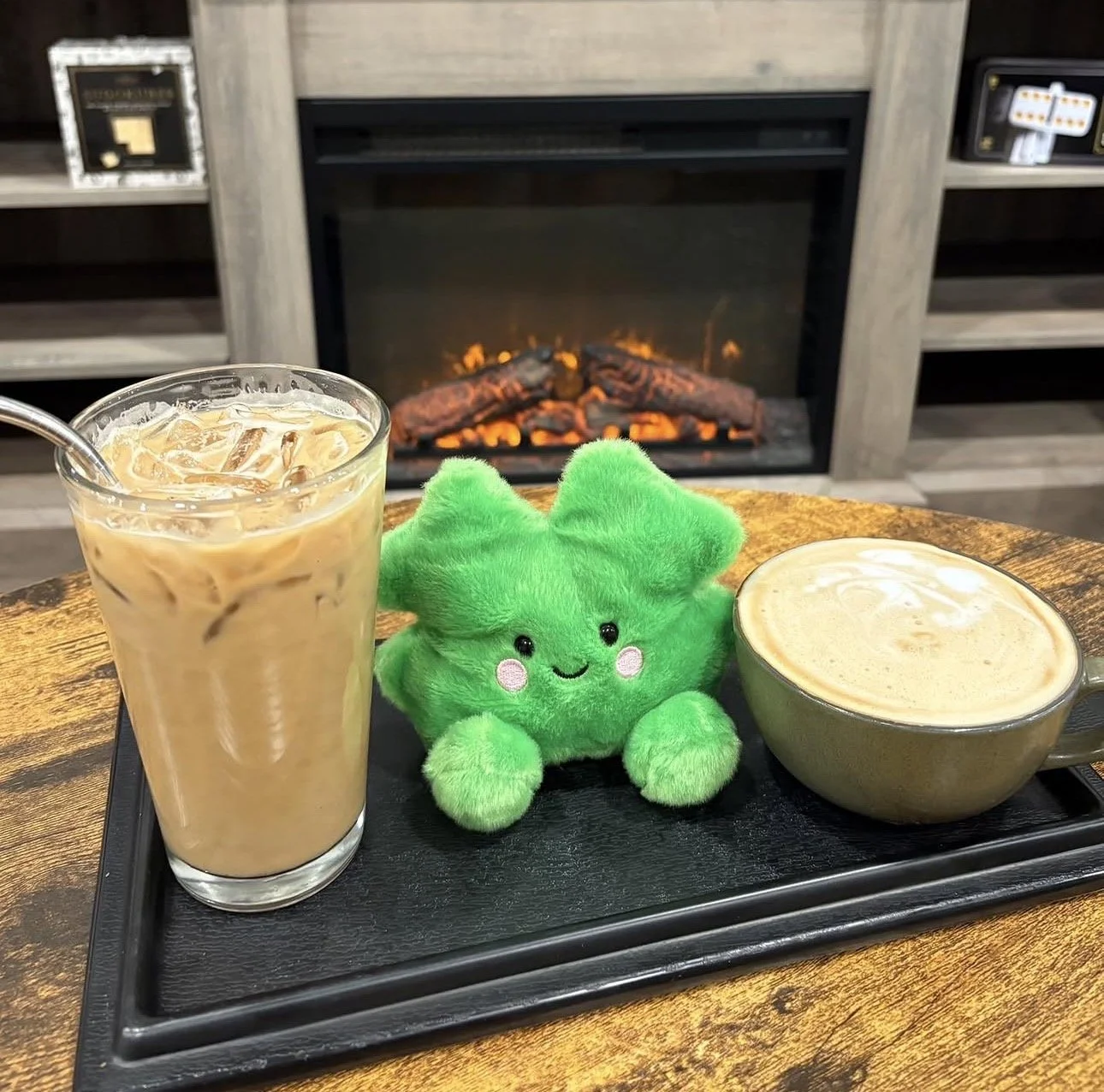 A tray with an iced coffee, a green plush cactus toy, and a cup of hot latte on a wooden table in front of a lit fireplace.