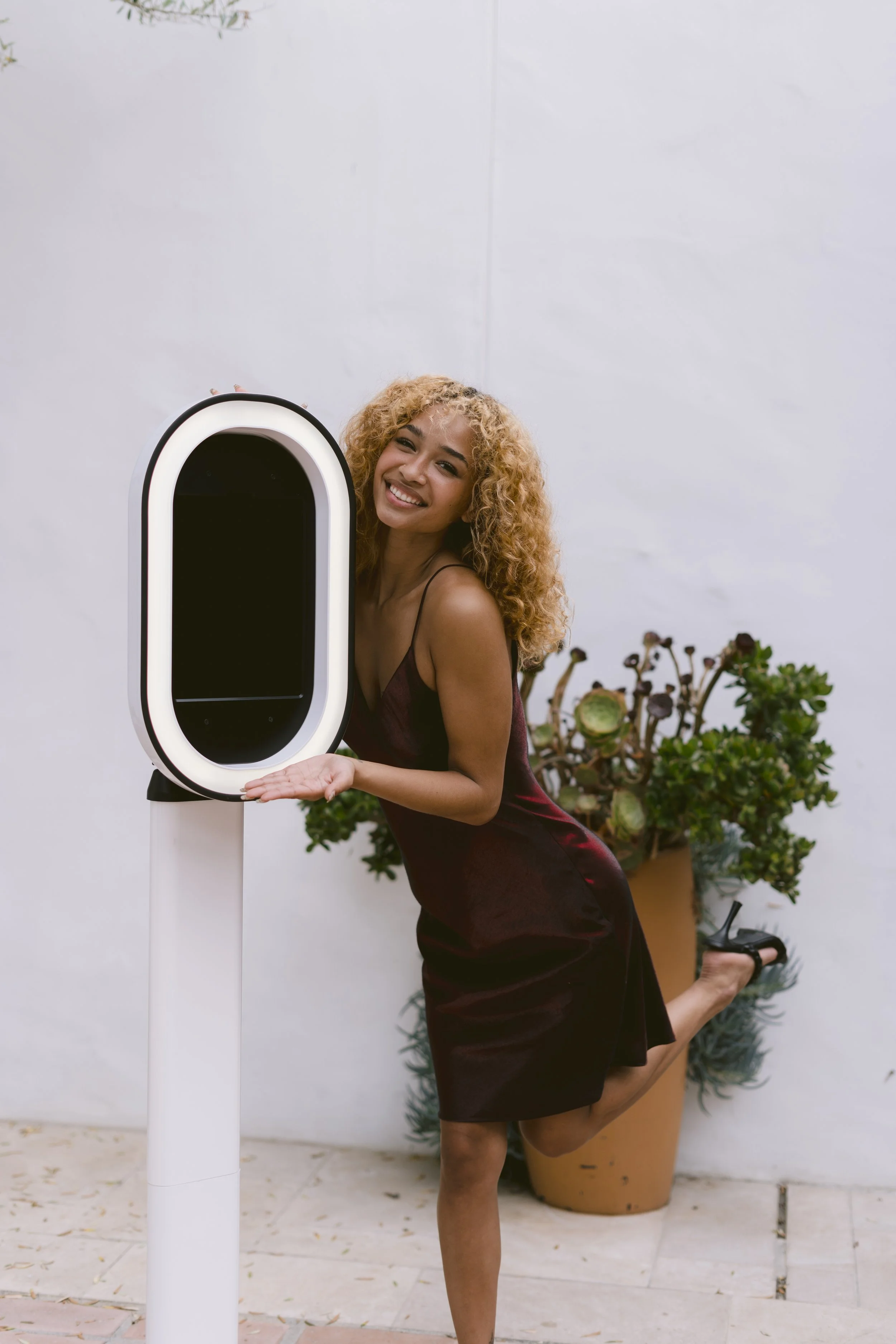 A woman with curly blonde hair is smiling, wearing a burgundy satin dress, and holding one leg up behind her while standing next to a black and white electric charging station outdoors, with potted plants in the background.