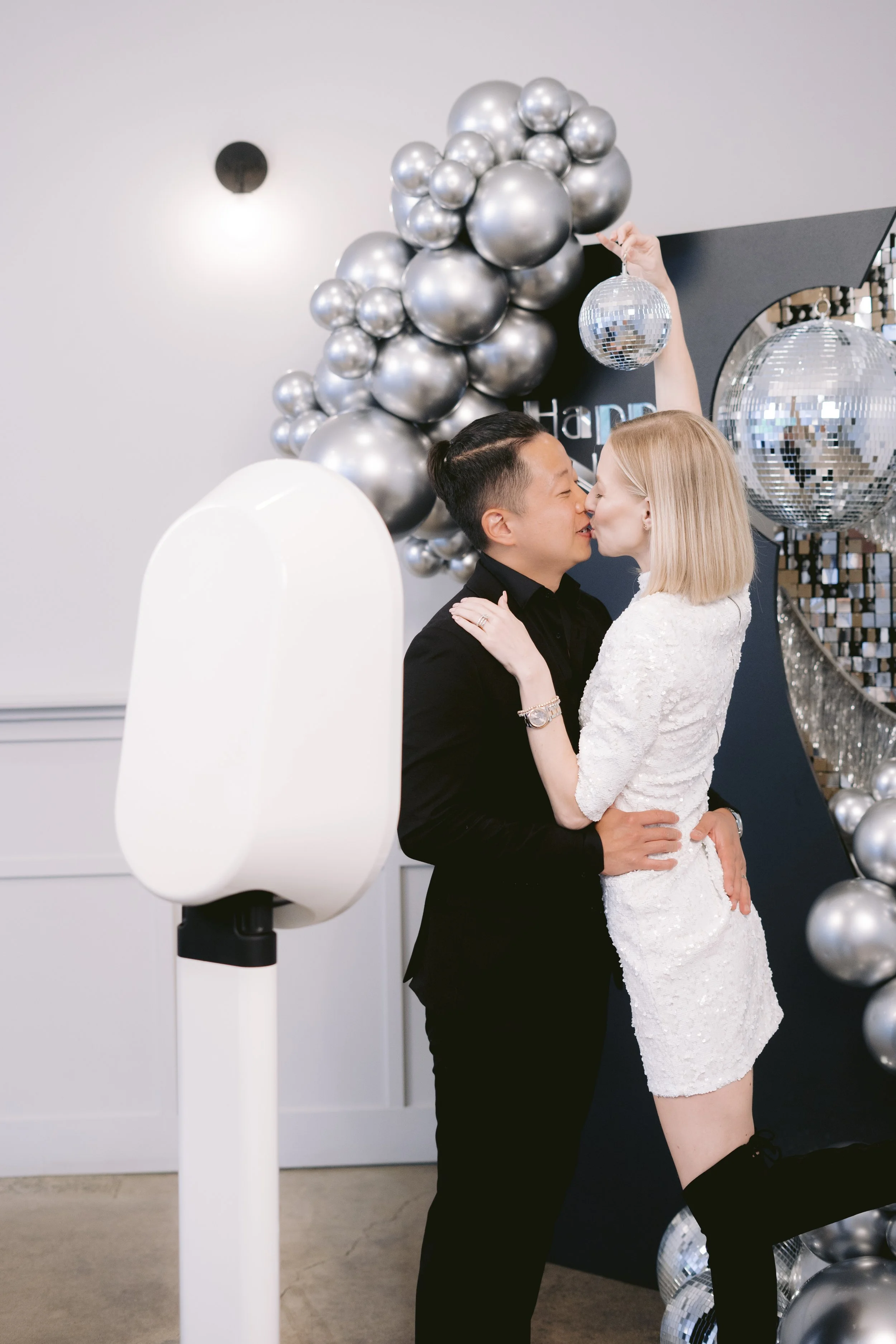 A couple is kissing at a celebration decorated with silver balloons and disco balls.