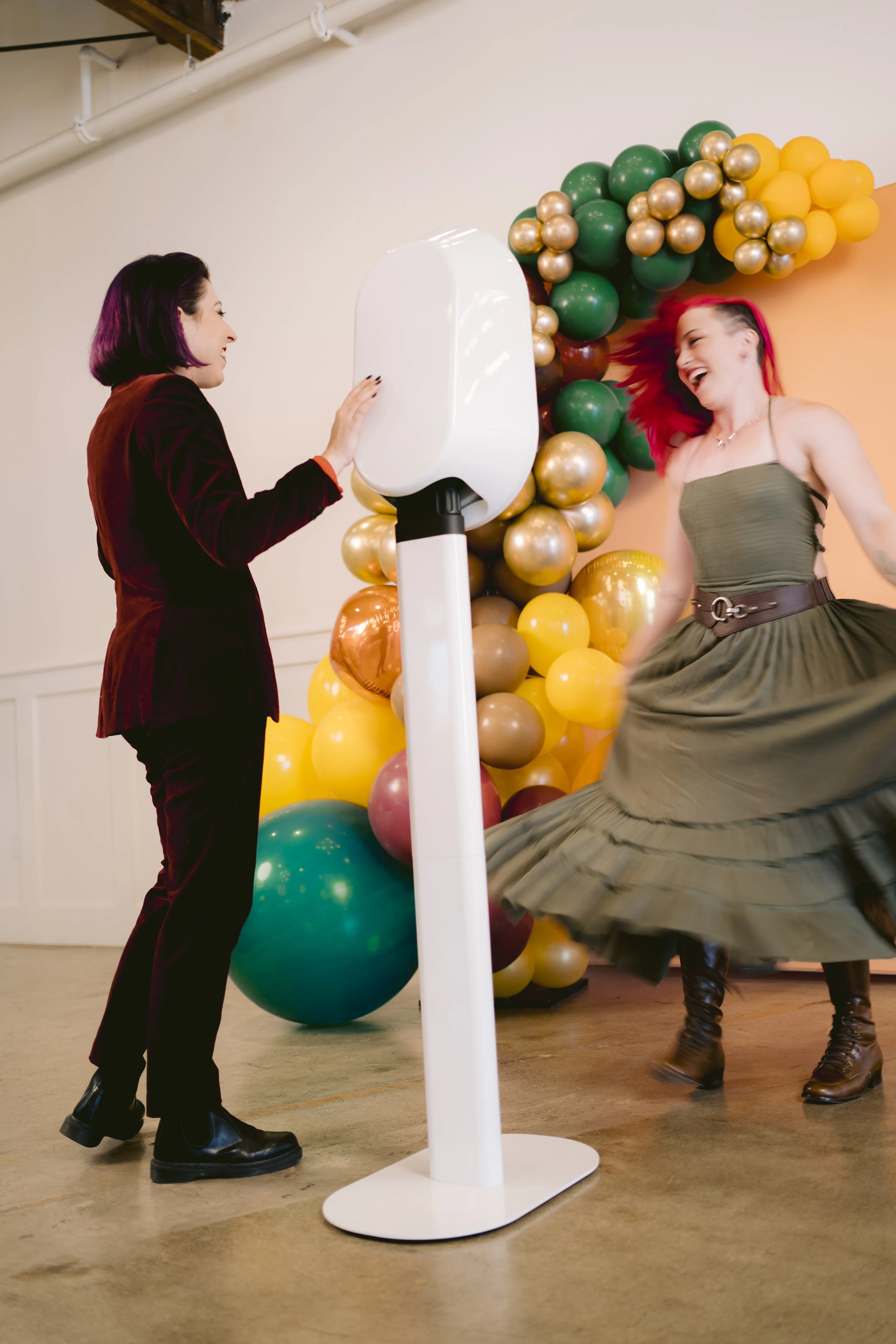 Two women, one in a dark red blazer and black boots, and the other in a green dress and brown boots, dancing and laughing in front of a balloon backdrop with gold, green, and yellow balloons.