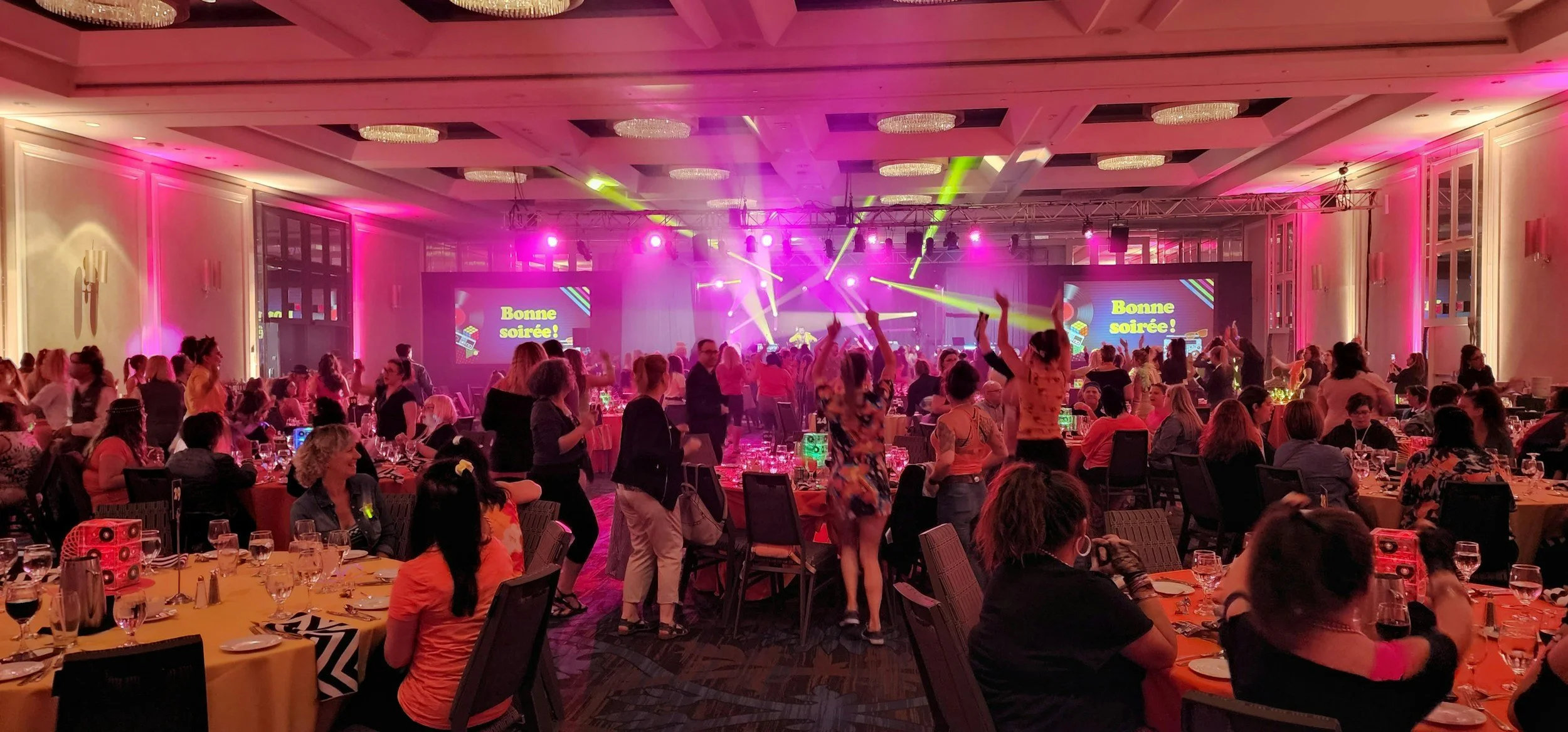 Two people talkinGuests dancing and dining at a corporate event with colorful stage lighting and branded screensg in a modern conference room with a stage and chairs, some attendees are seated in the background.