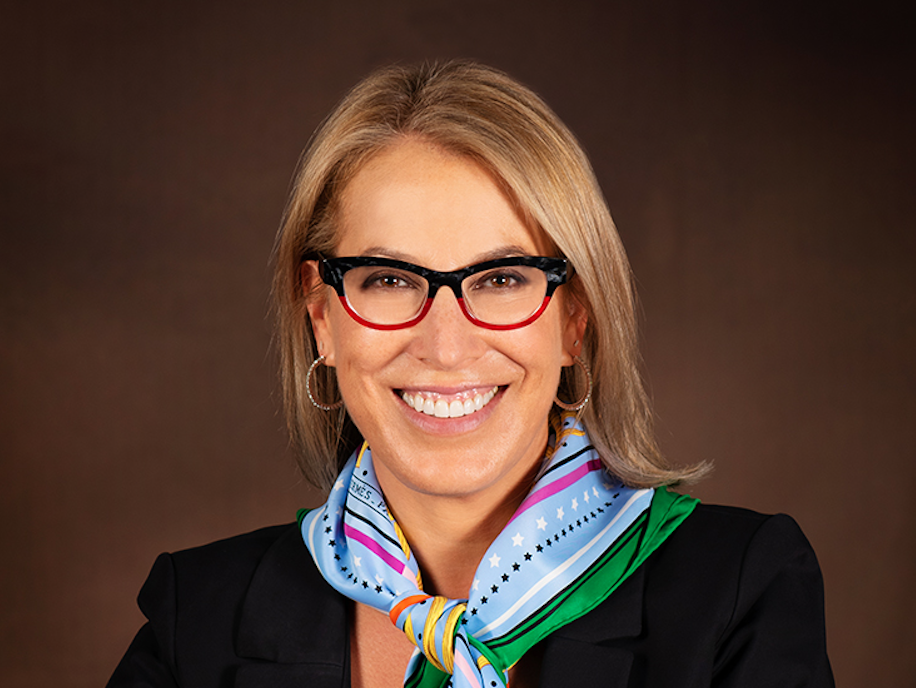 Lindsey Honari is a woman with blonde hair wearing glasses, a black blazer, and a colorful silk scarf, smiling at the camera against a plain brown background.