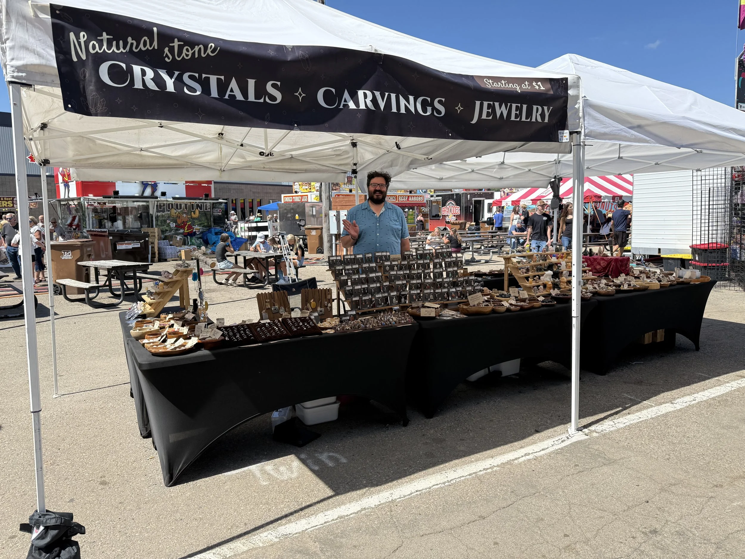 A vendor booth at an outdoor market or fair, selling crystals, carvings, jewelry, and other items. The booth has a black tablecloth and a large sign above it reading 'Natural stone, CRYSTALS, CARVINGS, JEWELRY' with a note indicating prices starting at $1. The background shows people walking and browsing other vendor tents and food stalls.
