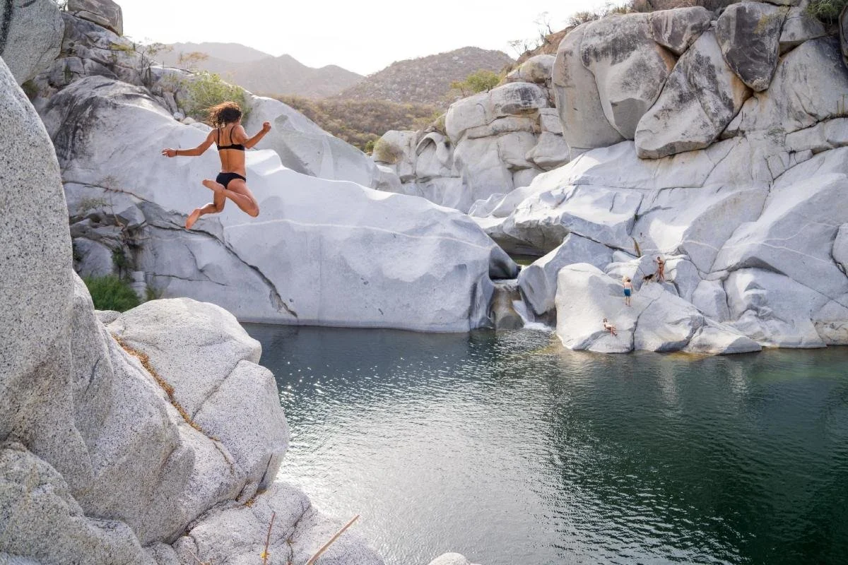 Person jumping off rock into natural pool, surrounded by large white boulders and hills in the background.