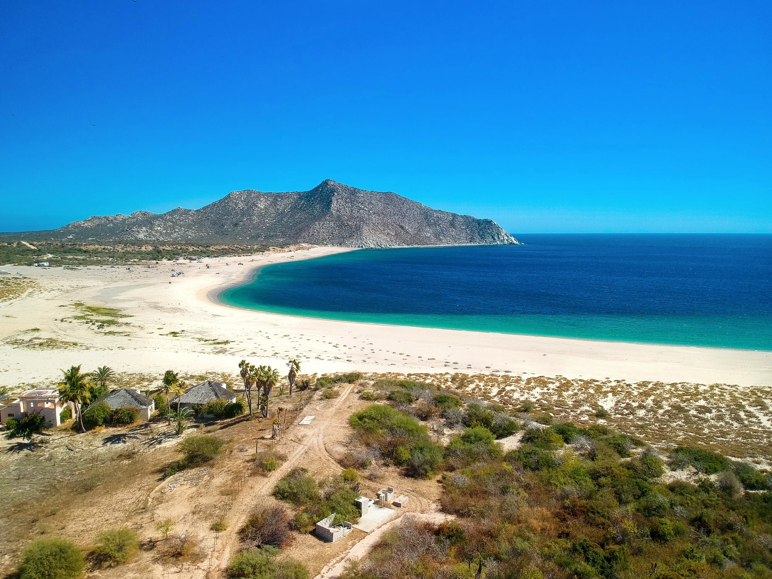 A scenic view of a coastal landscape with a sandy beach, turquoise ocean, and a distant mountain under a clear blue sky. Sparse bushes and small structures with thatched roofs are visible in the foreground.