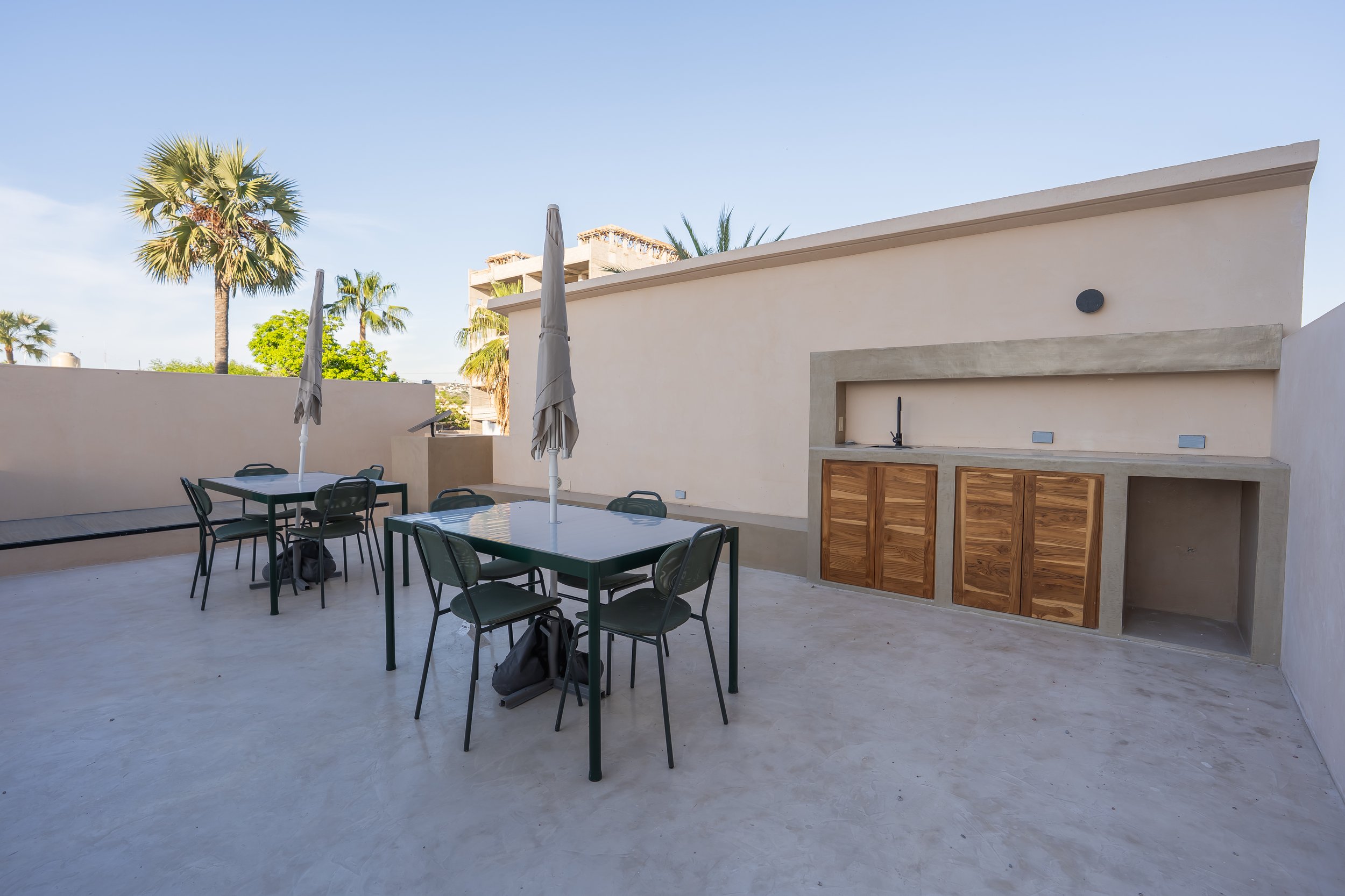 Outdoor rooftop patio with tables, chairs, umbrellas, palm trees, and a built-in barbecue area with wooden cabinets under a clear blue sky.