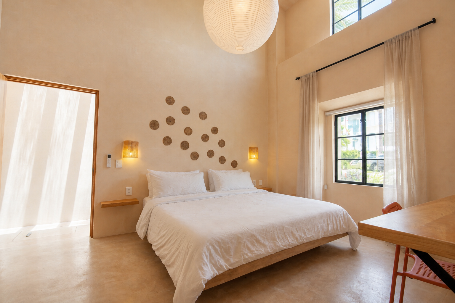 A cozy bedroom with a high ceiling, large white bed, and natural light from tall windows. The walls are beige with circular wall decor, and there are warm wall-mounted lamps on either side of the bed.