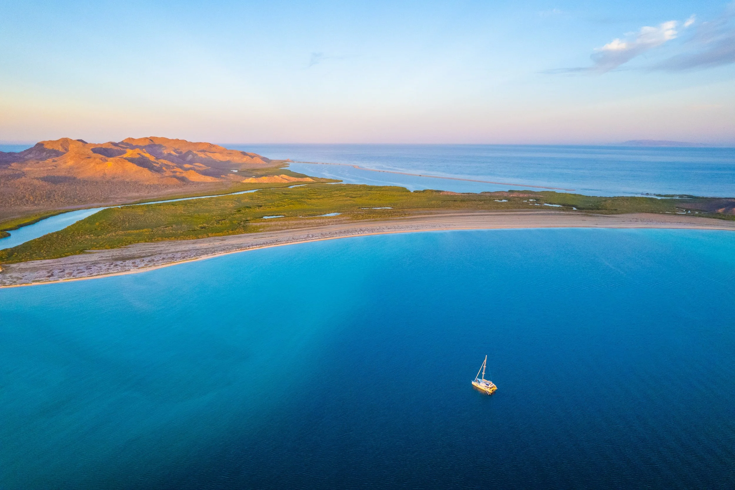 Aerial view of a coastal landscape with mountains, green wetlands, and a large body of water with a sailboat.
