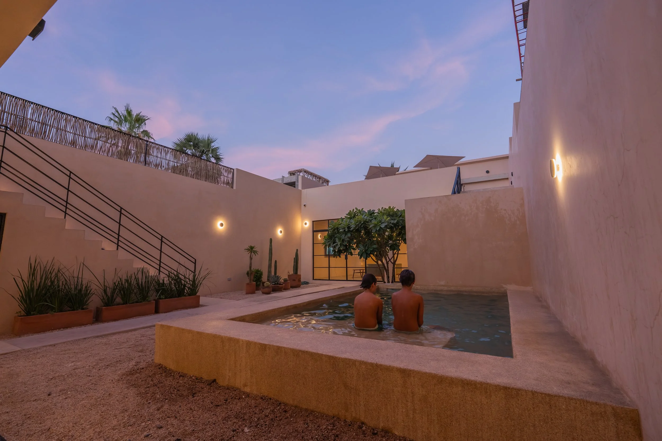 Two people sitting in a swimming pool at sunset in a modern courtyard with beige walls, potted plants, and outdoor lighting.
